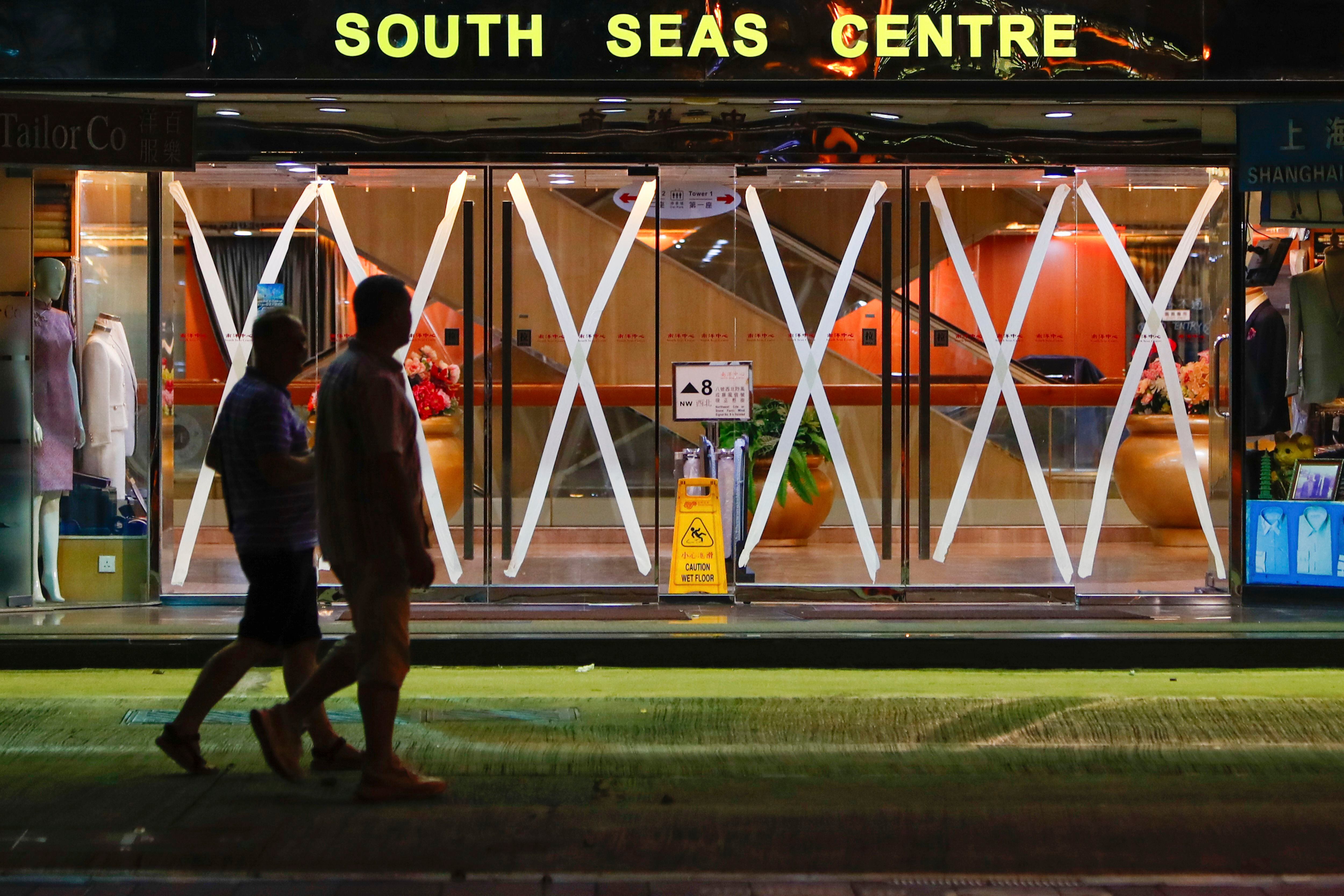 Dos personas pasan junto a un centro comercial cuyas puertas de vidrio tienen tiras de cinta, en precaución contra el tifón Saola que se acerca, el viernes 1 de septiembre de 2023, en Hong Kong. (AP Foto/Daniel Ceng)