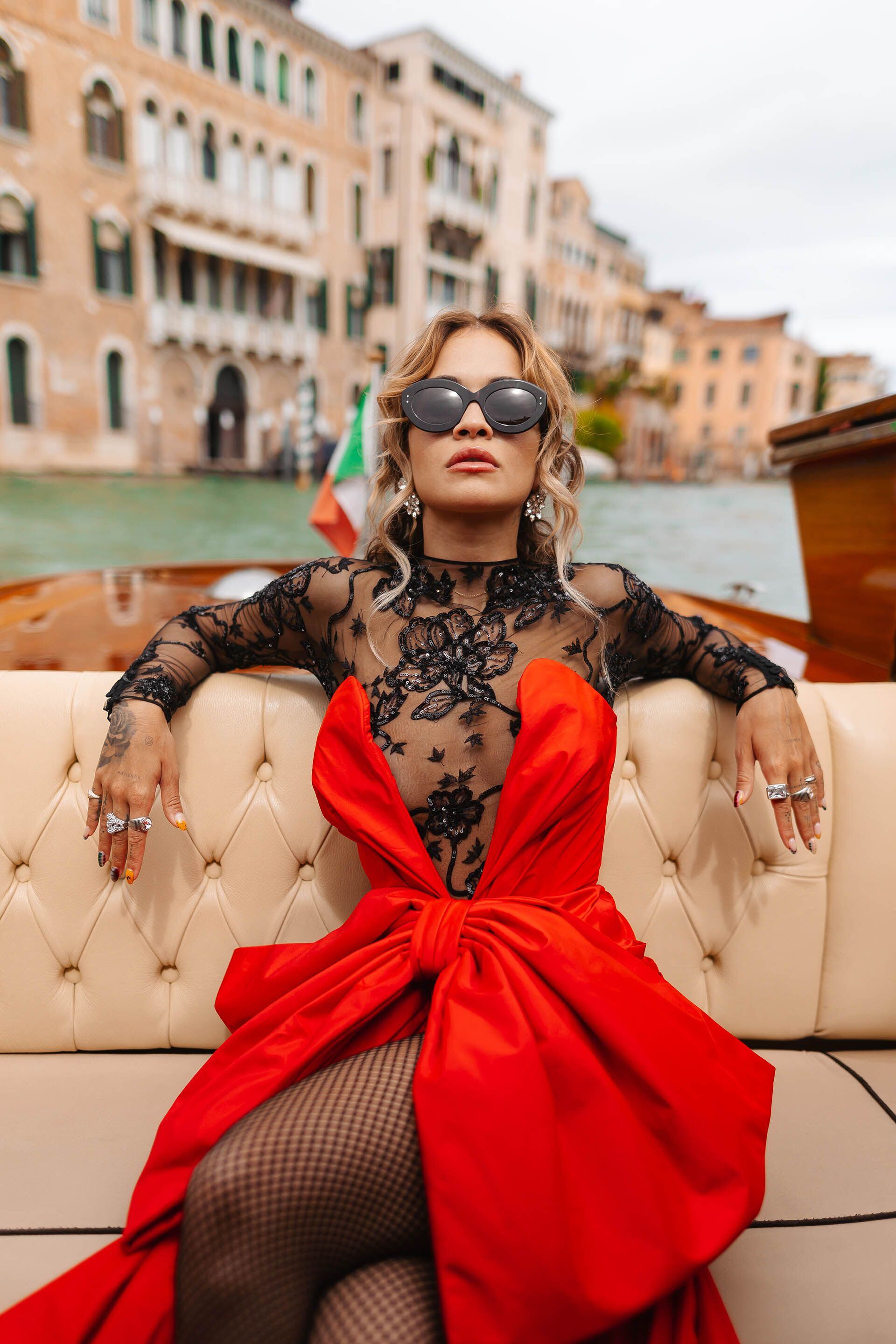 Rita Ora realizó una producción en una embarcación en Venecia, Italia, a donde viajó para ser parte del Festival de Cine. La artista lució un vestido de Moschino que combinó encaje negro y un moño grande color rojo