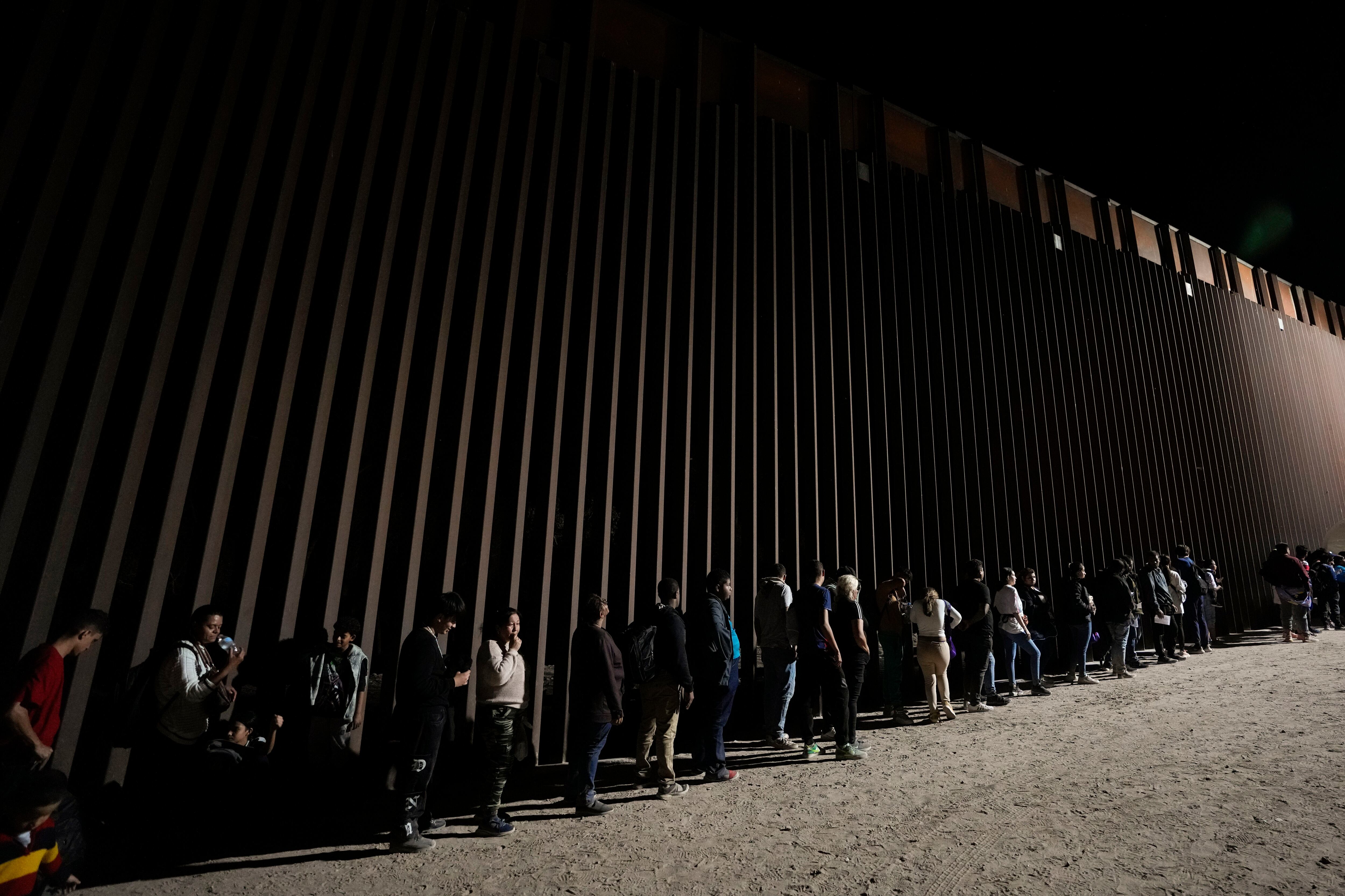 Un grupo de personas hacen fila junto al muro fronterizo mientras esperan para solicitar asilo tras cruzar al frontera desde México. (AP Foto/Gregory Bull)