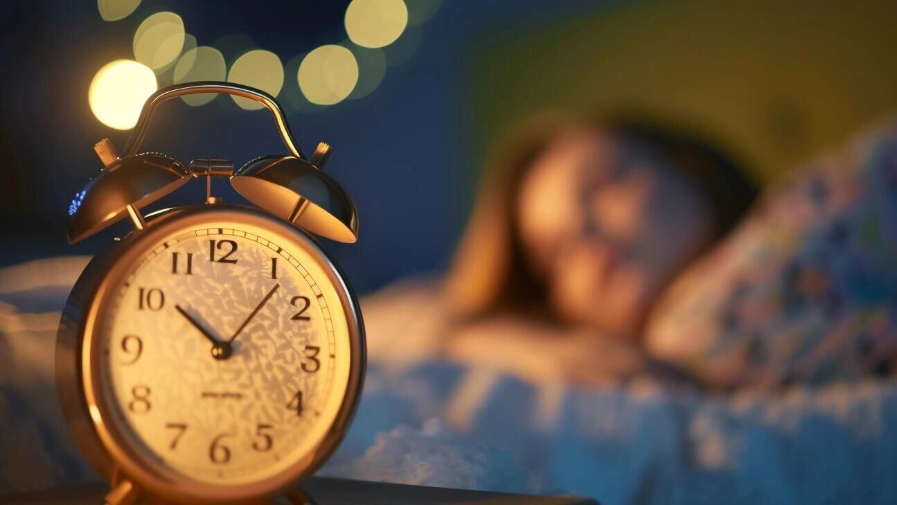 En esta escena nocturna, una mujer duerme serenamente en su cama, con un reloj despertador enfocado en primer plano, preparado para iniciar el día. La imagen enfatiza el valor del descanso reparador y la función del sueño en la promoción de una buena salud mental, ilustrando la conexión entre dormir bien y sentirse bien. (Imagen ilustrativa Infobae)