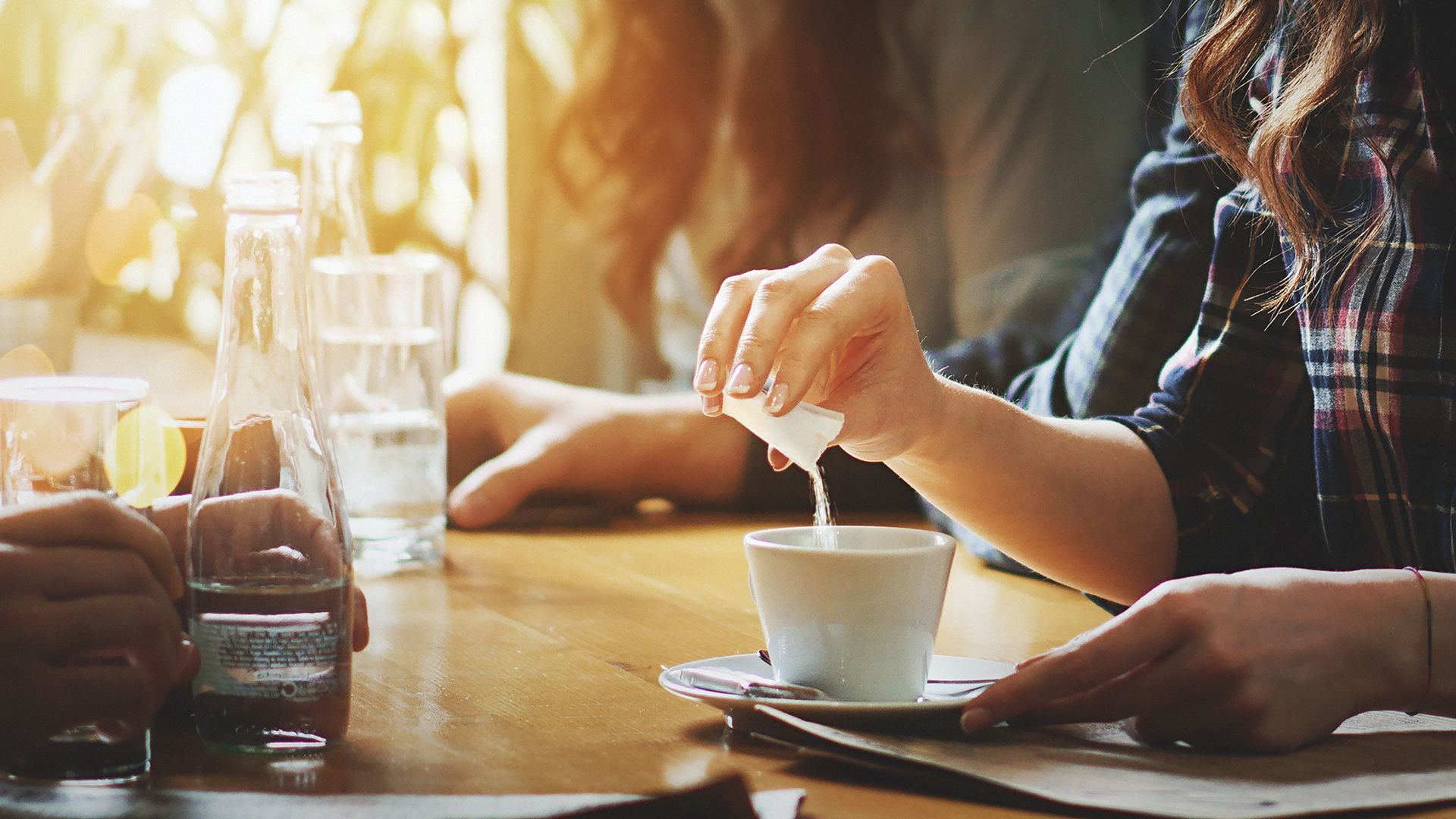 Closeup of unrecognizable woman putting some sugar into her coffee. She's having a tea spoon of sugar packed in a little bag. Blurry people in background.