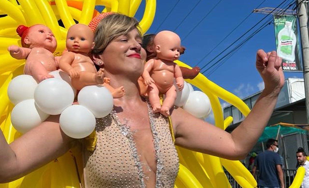 Woman in costume. Three baby dolls are sewn on his shoulders. His back has long yellow balloons.