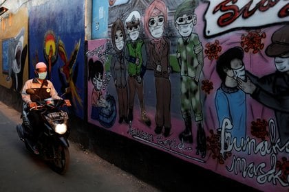 Un hombre con una máscara protectora conduce una motocicleta junto a murales que promueven la conciencia de la enfermedad por coronavirus en un callejón de una aldea en Bekasi, en las afueras de Yakarta, Indonesia (Reuters)