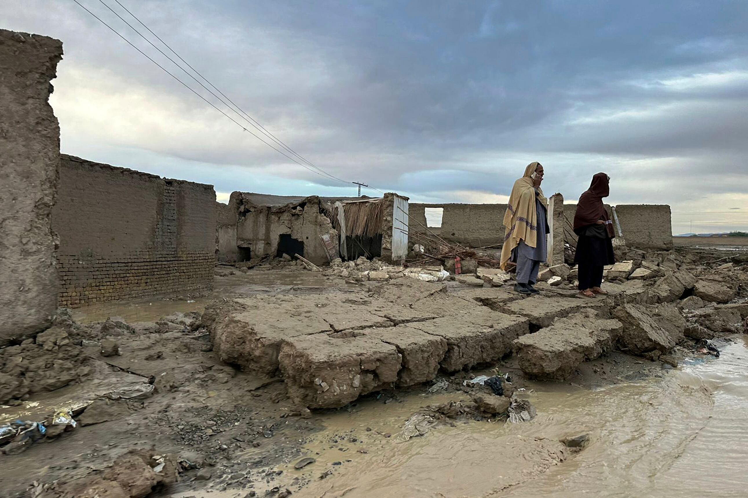 Personas permanecen de pie sobre un muro caído de su vivienda, que sufrió daños debido a las fuertes lluvias en un área cerca de Chaman, Pakistán, el jueves 18 de abril de 2024 (AP Foto/Habib Ullah)