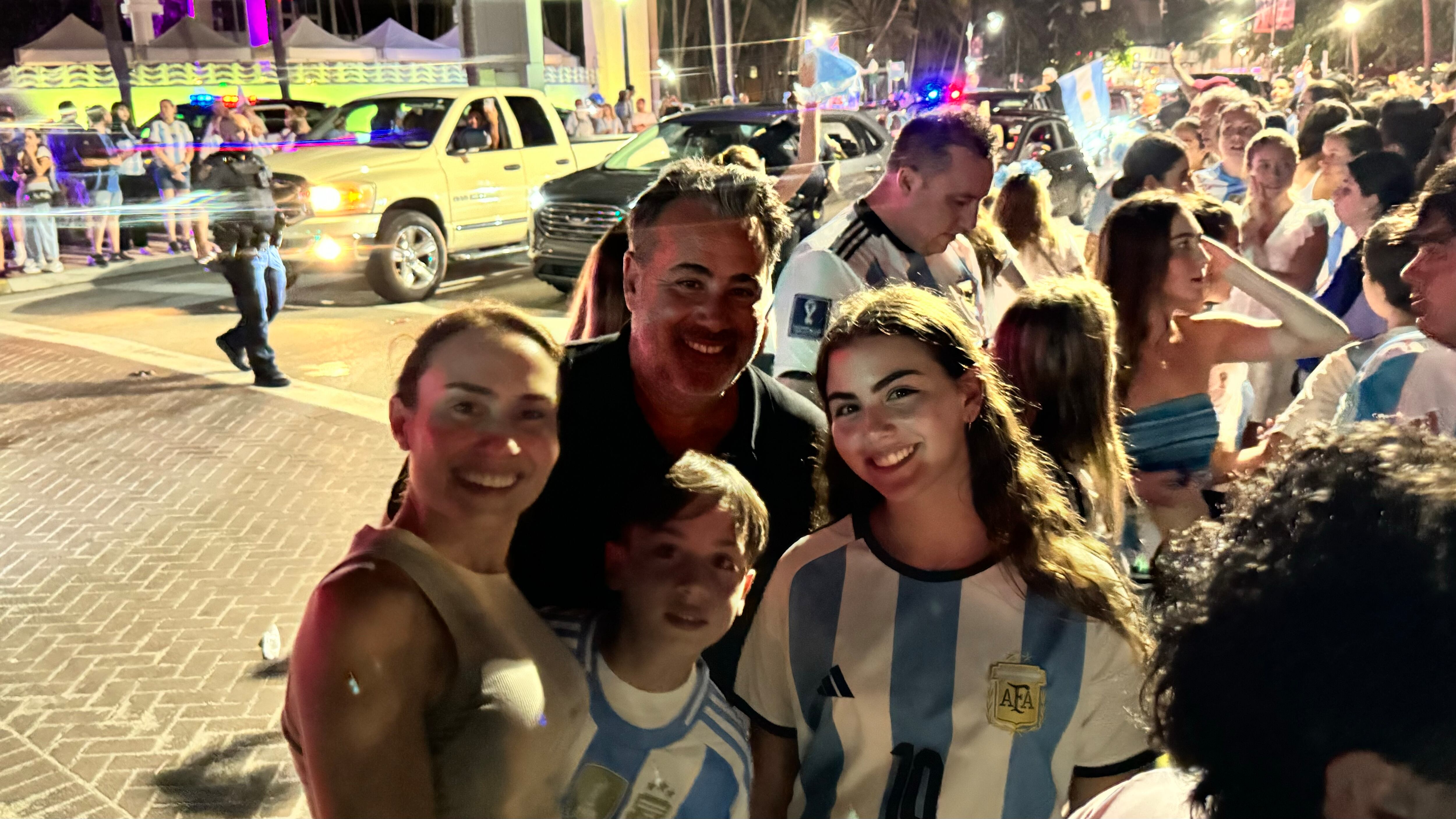 Una multitud de hinchas argentinos, vestidos con camisetas de la selección y agitando banderas celestes y blancas, celebra en las calles de Miami Beach durante la noche. La gente se muestra eufórica, tomando fotos y cantando, en una atmósfera festiva y llena de emoción tras la victoria de la Selección Argentina en la Copa América.