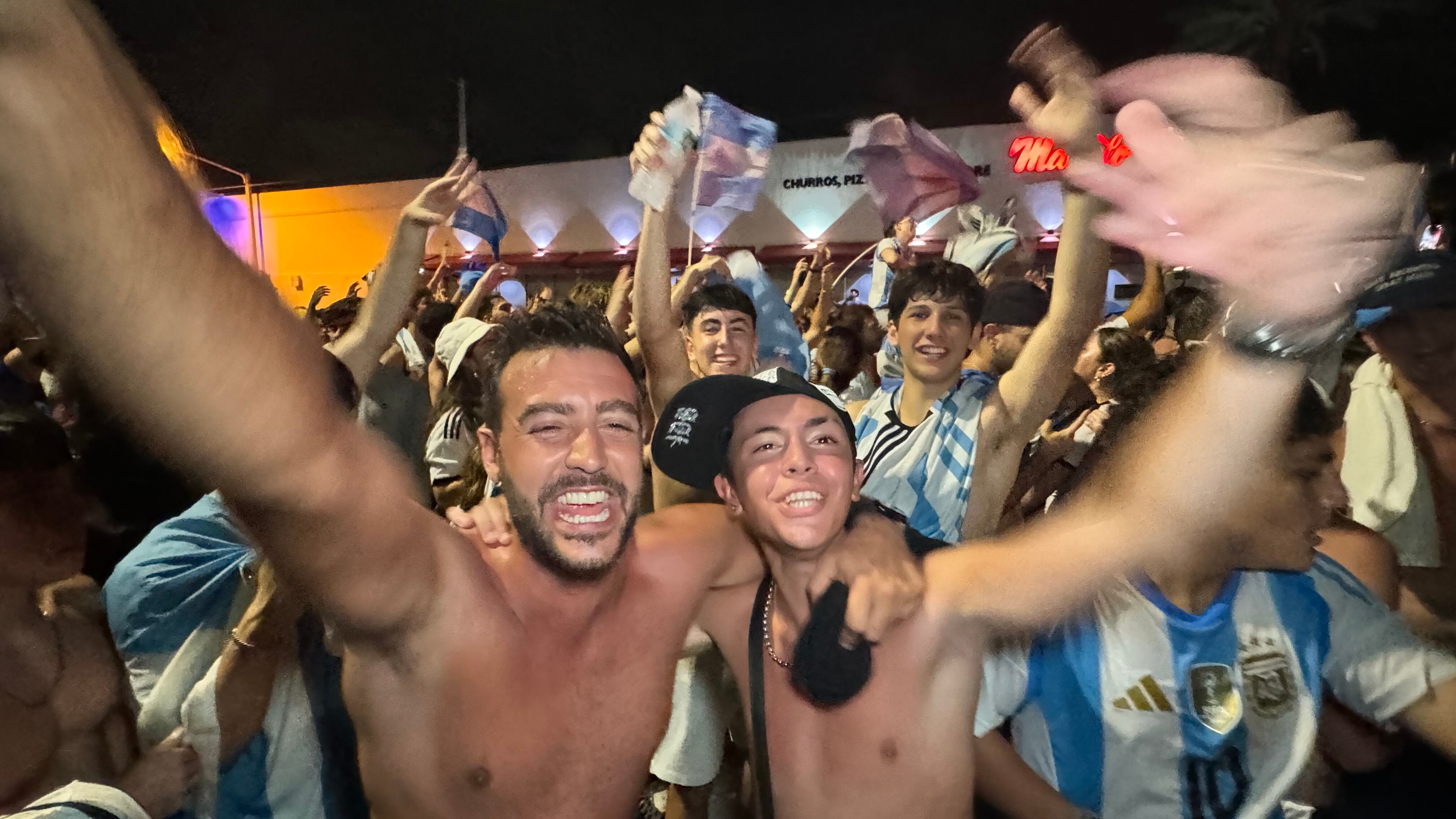 Una multitud de hinchas argentinos, vestidos con camisetas de la selección y agitando banderas celestes y blancas, celebra en las calles de Miami Beach durante la noche. La gente se muestra eufórica, tomando fotos y cantando, en una atmósfera festiva y llena de emoción tras la victoria de la Selección Argentina en la Copa América.