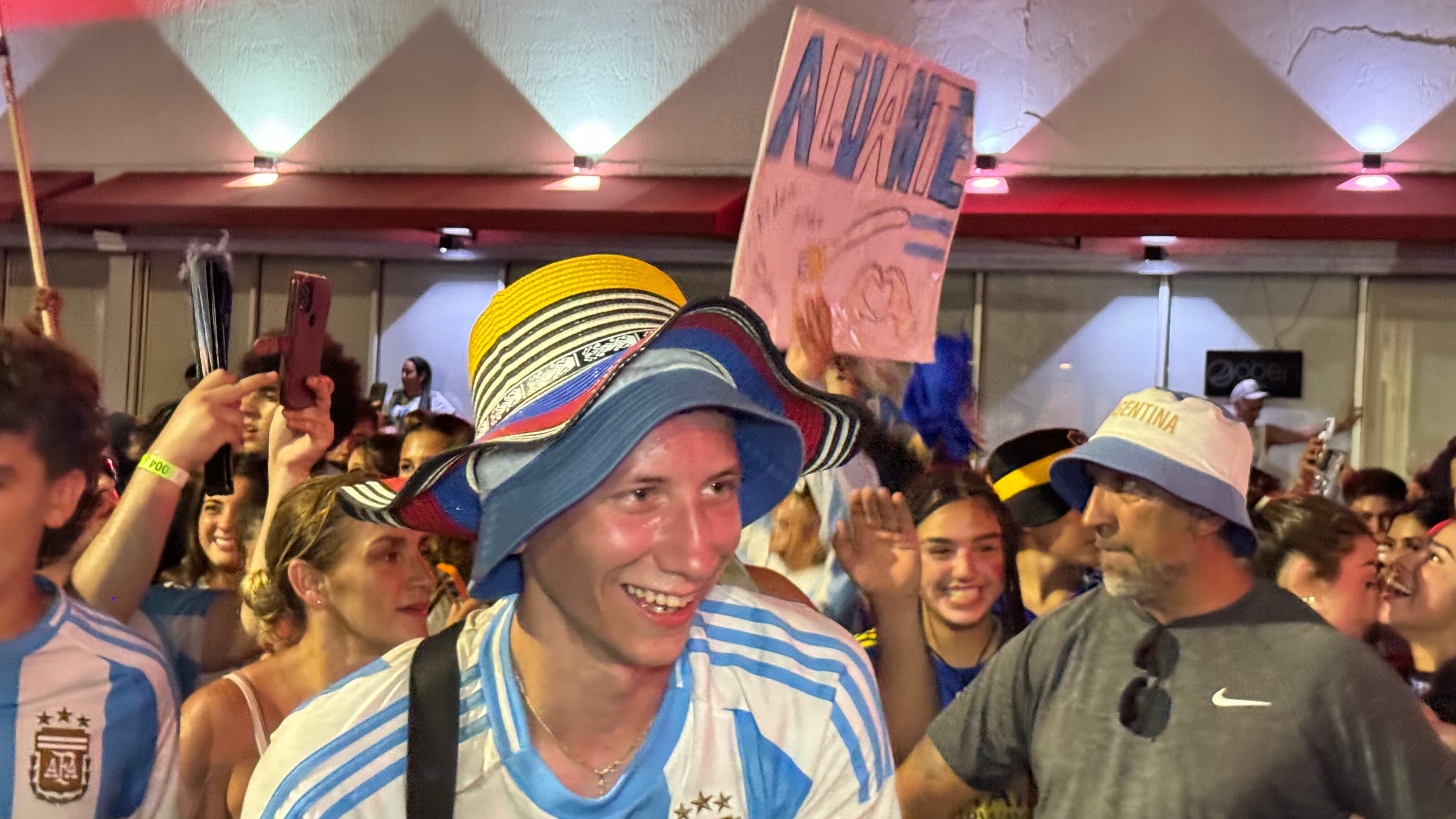 Una multitud de hinchas argentinos, vestidos con camisetas de la selección y agitando banderas celestes y blancas, celebra en las calles de Miami Beach durante la noche. La gente se muestra eufórica, tomando fotos y cantando, en una atmósfera festiva y llena de emoción tras la victoria de la Selección Argentina en la Copa América.