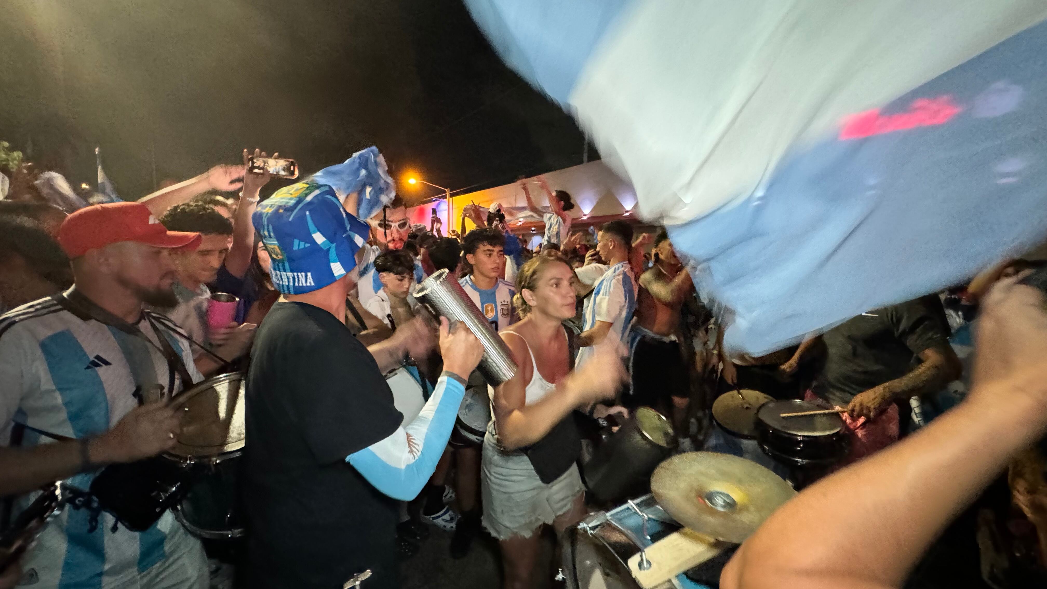 Una multitud de hinchas argentinos, vestidos con camisetas de la selección y agitando banderas celestes y blancas, celebra en las calles de Miami Beach durante la noche. La gente se muestra eufórica, tomando fotos y cantando, en una atmósfera festiva y llena de emoción tras la victoria de la Selección Argentina en la Copa América.