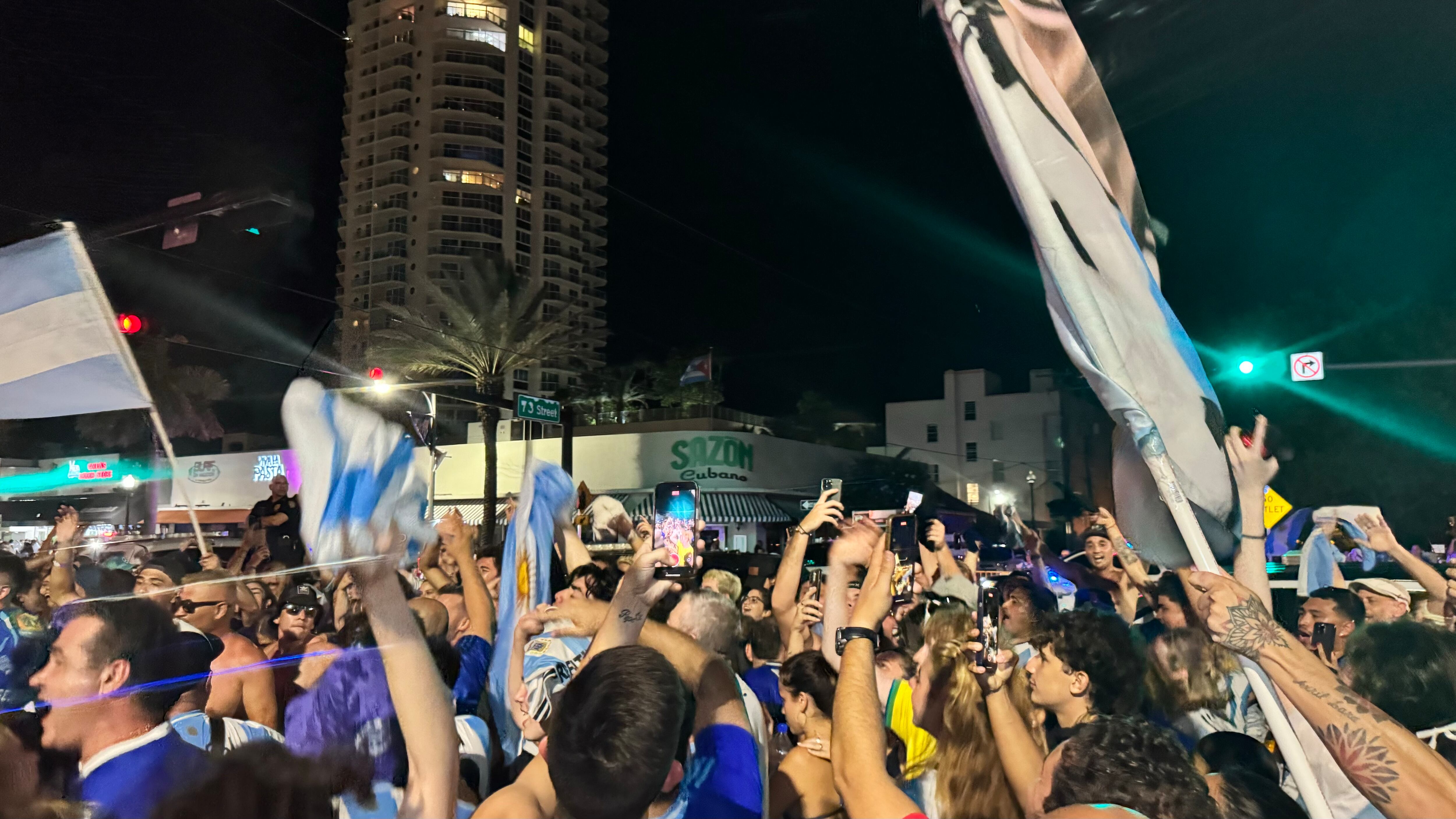 Una multitud de hinchas argentinos, vestidos con camisetas de la selección y agitando banderas celestes y blancas, celebra en las calles de Miami Beach durante la noche. La gente se muestra eufórica, tomando fotos y cantando, en una atmósfera festiva y llena de emoción tras la victoria de la Selección Argentina en la Copa América.