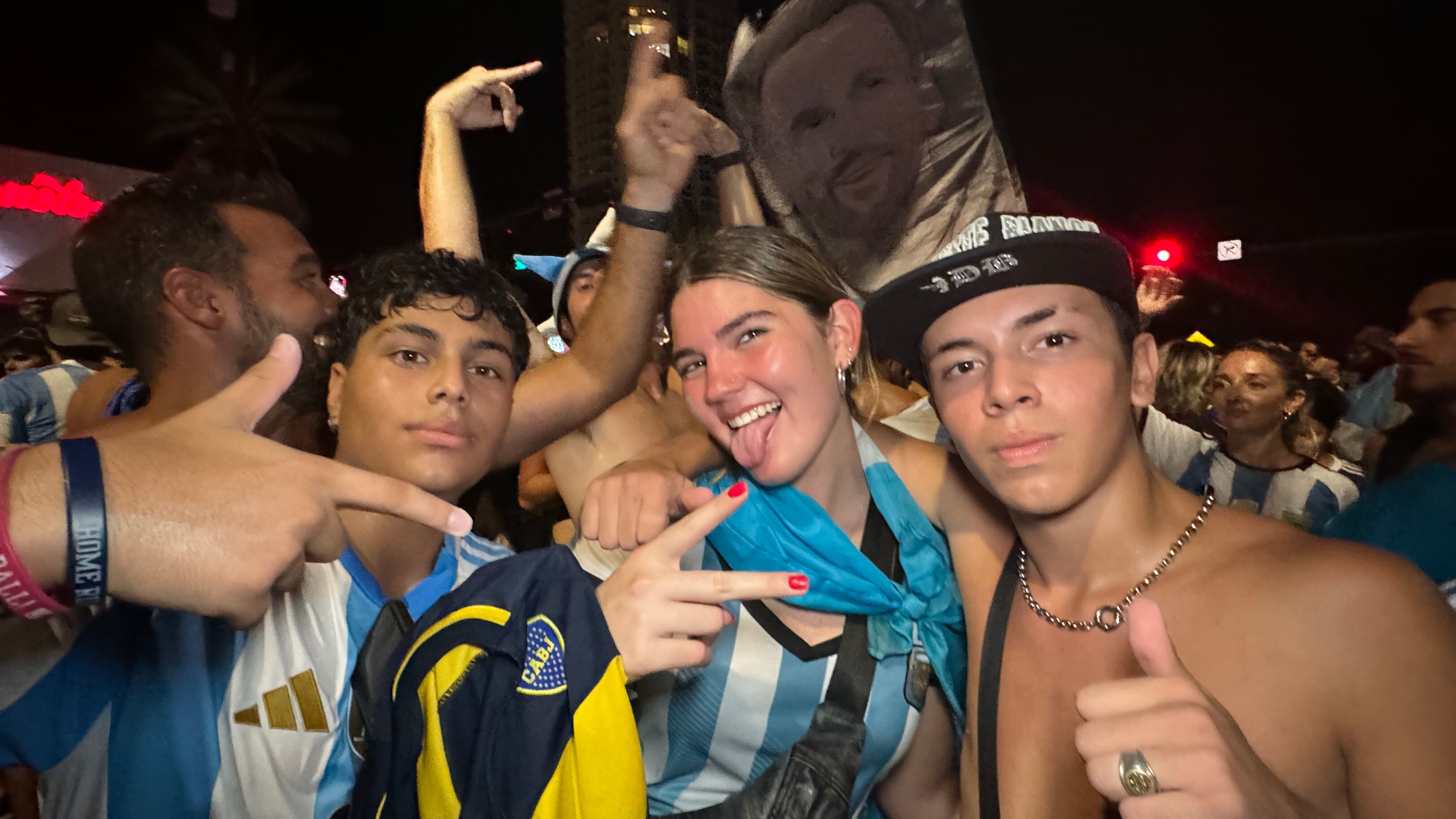 Una multitud de hinchas argentinos, vestidos con camisetas de la selección y agitando banderas celestes y blancas, celebra en las calles de Miami Beach durante la noche. La gente se muestra eufórica, tomando fotos y cantando, en una atmósfera festiva y llena de emoción tras la victoria de la Selección Argentina en la Copa América.