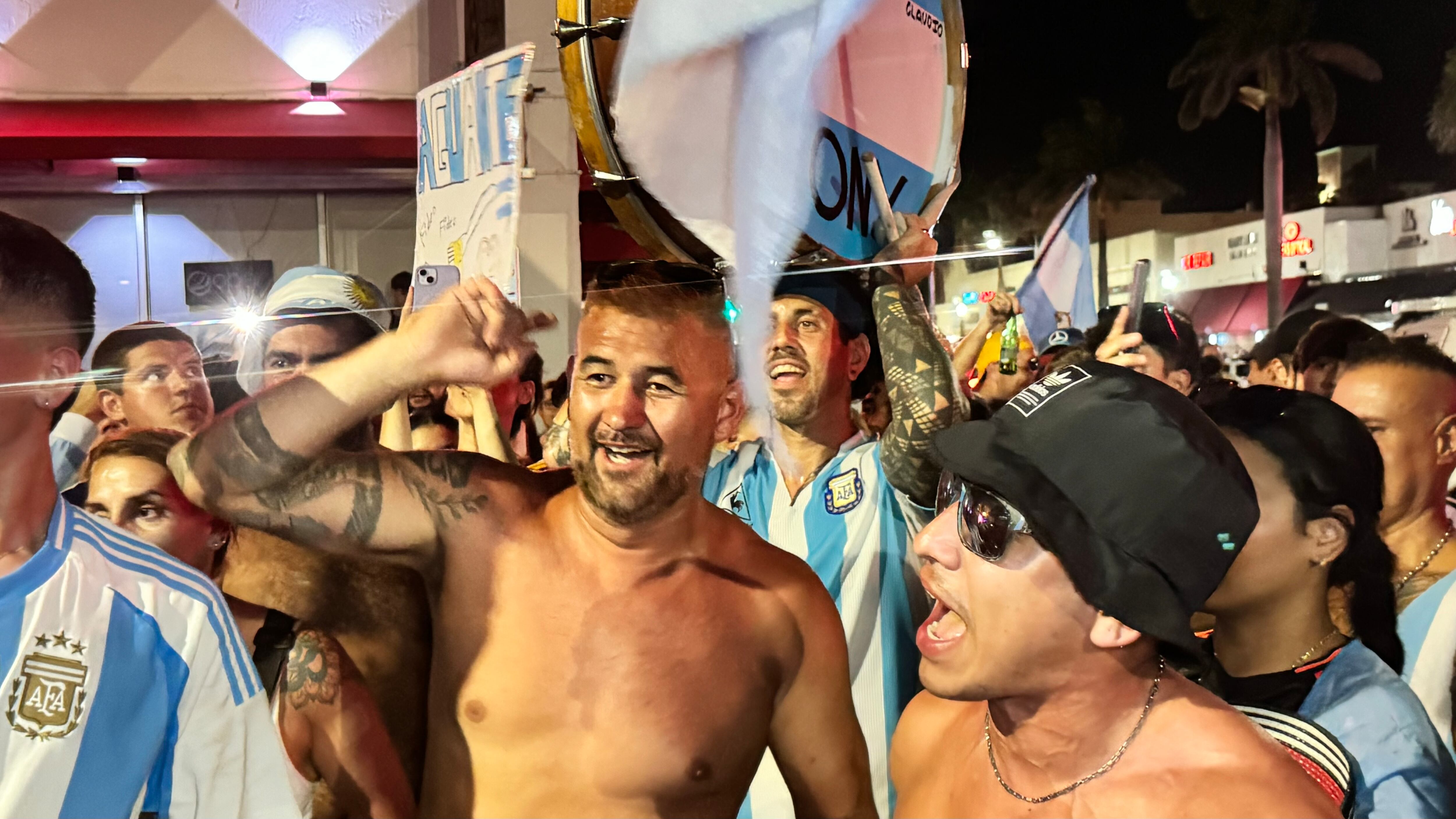 Una multitud de hinchas argentinos, vestidos con camisetas de la selección y agitando banderas celestes y blancas, celebra en las calles de Miami Beach durante la noche. La gente se muestra eufórica, tomando fotos y cantando, en una atmósfera festiva y llena de emoción tras la victoria de la Selección Argentina en la Copa América.