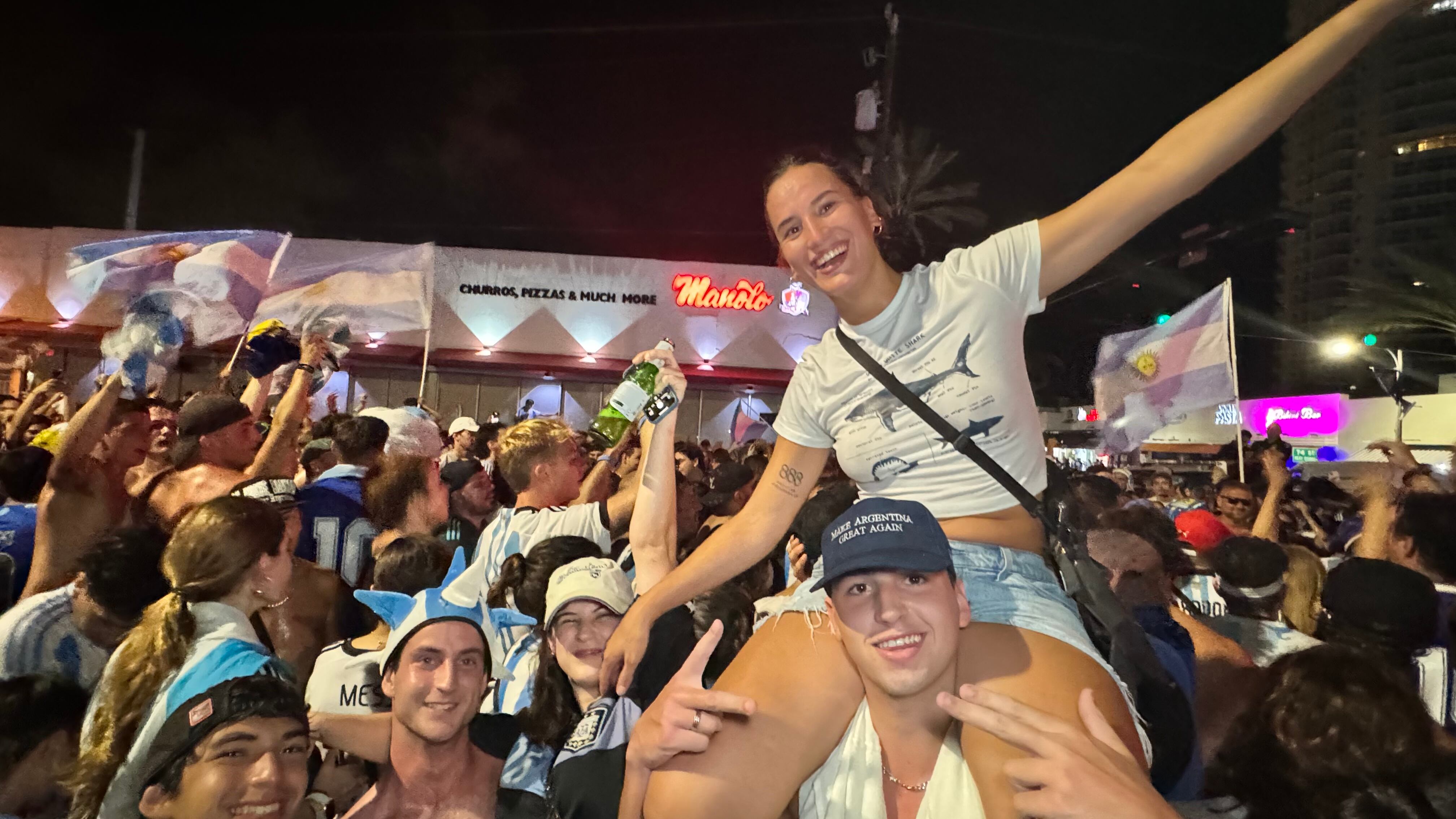 Una multitud de hinchas argentinos, vestidos con camisetas de la selección y agitando banderas celestes y blancas, celebra en las calles de Miami Beach durante la noche. La gente se muestra eufórica, tomando fotos y cantando, en una atmósfera festiva y llena de emoción tras la victoria de la Selección Argentina en la Copa América.