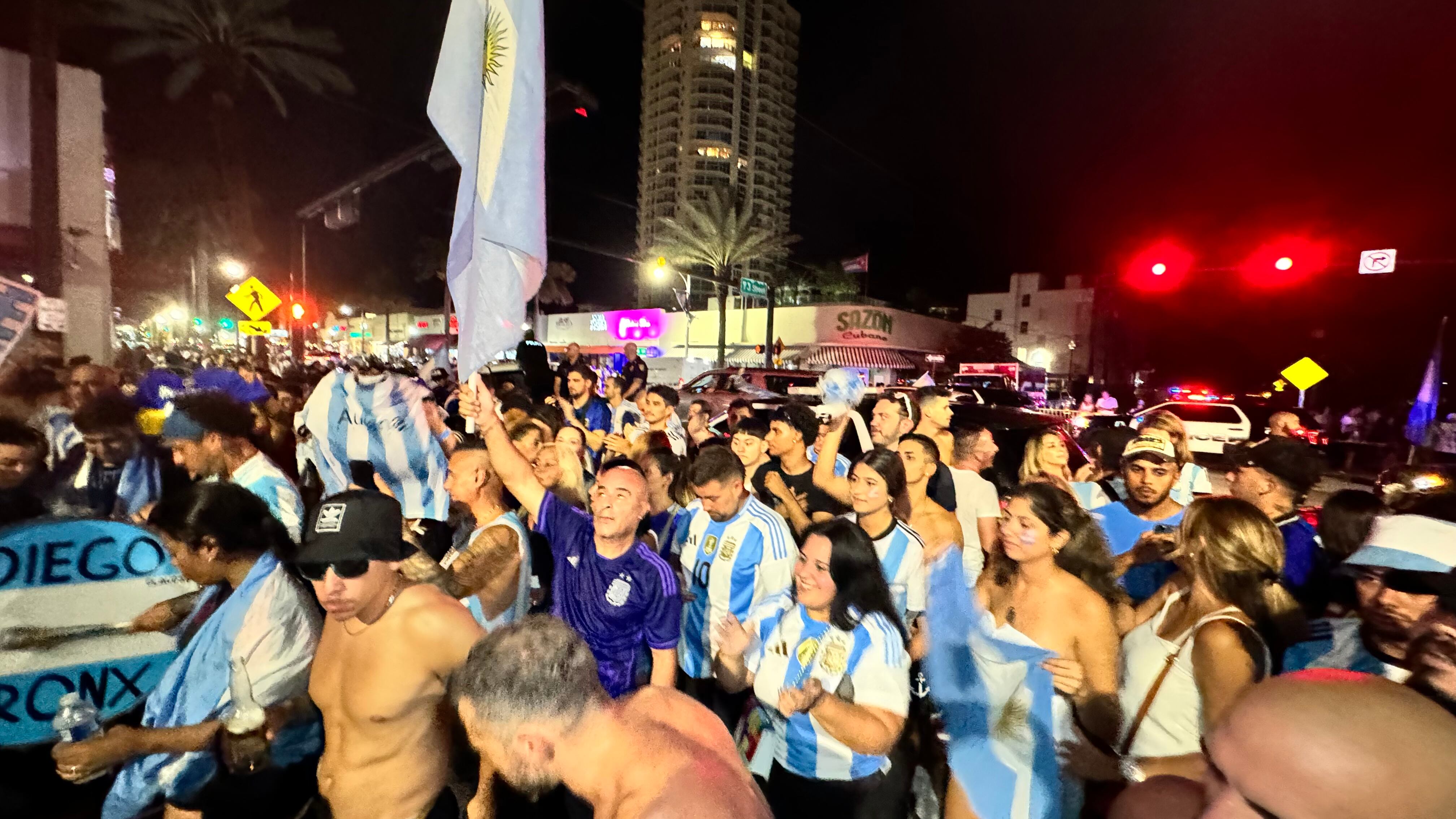 Una multitud de hinchas argentinos, vestidos con camisetas de la selección y agitando banderas celestes y blancas, celebra en las calles de Miami Beach durante la noche. La gente se muestra eufórica, tomando fotos y cantando, en una atmósfera festiva y llena de emoción tras la victoria de la Selección Argentina en la Copa América.