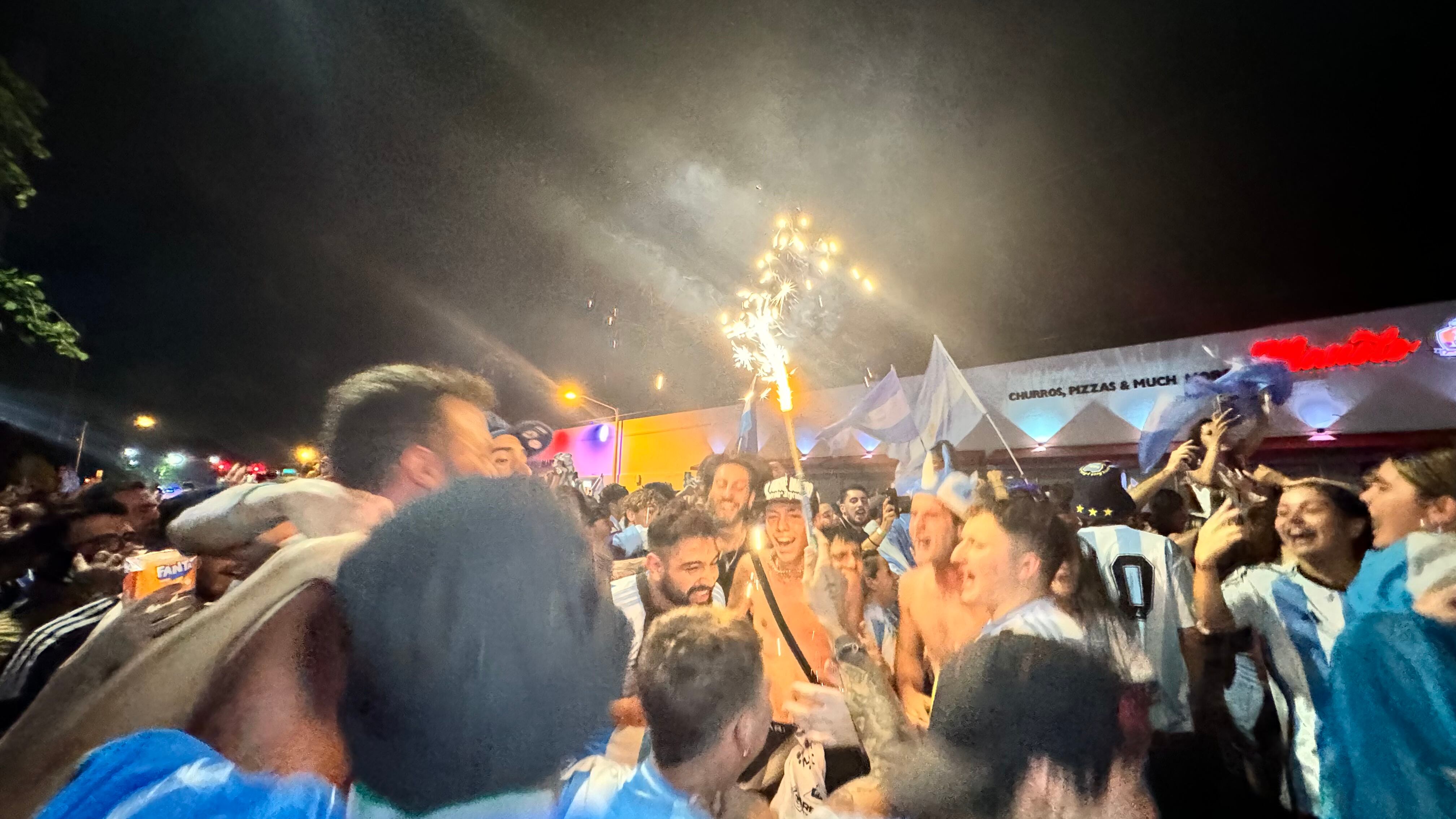 Una multitud de hinchas argentinos, vestidos con camisetas de la selección y agitando banderas celestes y blancas, celebra en las calles de Miami Beach durante la noche. La gente se muestra eufórica, tomando fotos y cantando, en una atmósfera festiva y llena de emoción tras la victoria de la Selección Argentina en la Copa América.