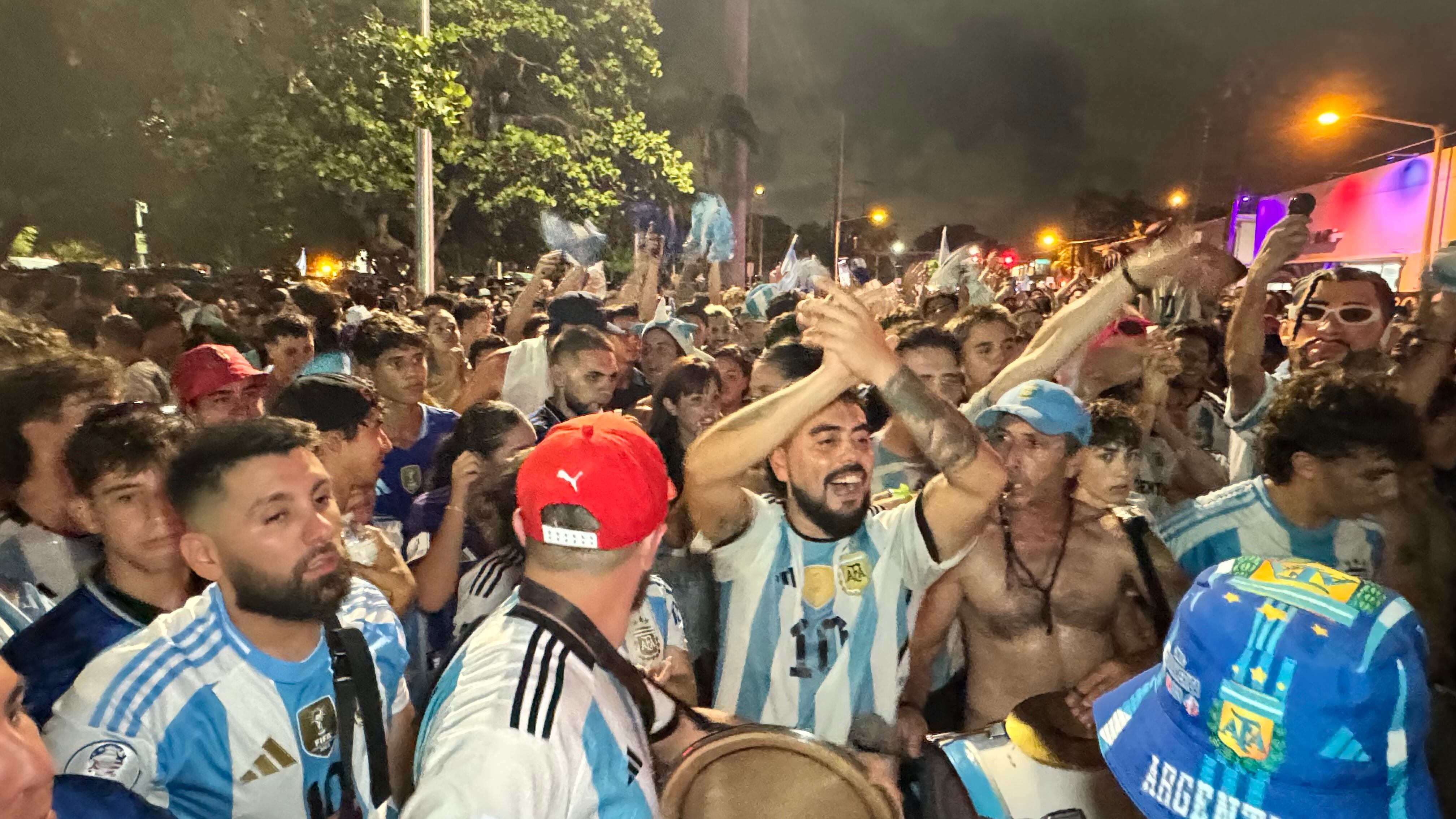 Una multitud de hinchas argentinos, vestidos con camisetas de la selección y agitando banderas celestes y blancas, celebra en las calles de Miami Beach durante la noche. La gente se muestra eufórica, tomando fotos y cantando, en una atmósfera festiva y llena de emoción tras la victoria de la Selección Argentina en la Copa América.