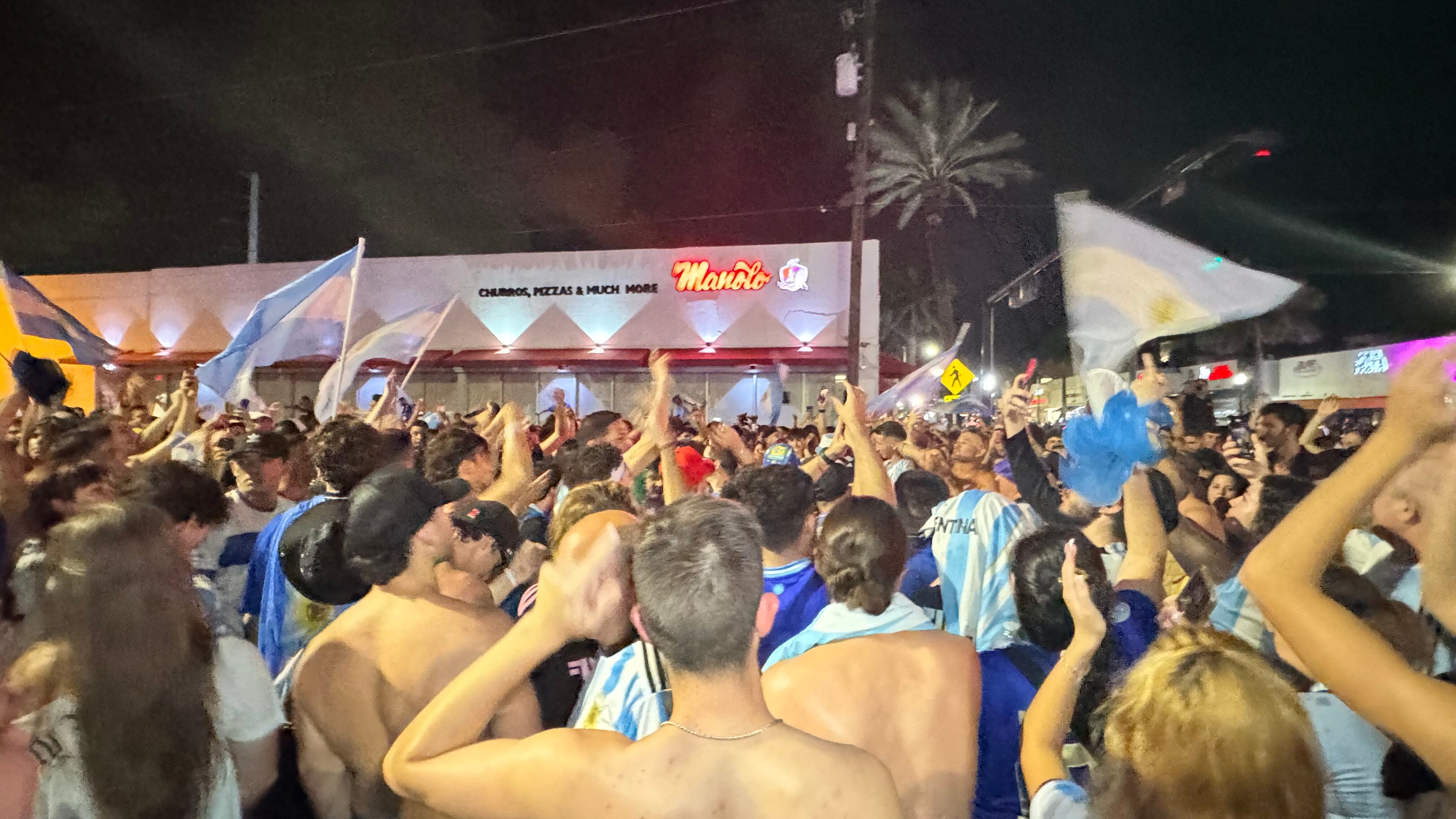 Una multitud de hinchas argentinos, vestidos con camisetas de la selección y agitando banderas celestes y blancas, celebra en las calles de Miami Beach durante la noche. La gente se muestra eufórica, tomando fotos y cantando, en una atmósfera festiva y llena de emoción tras la victoria de la Selección Argentina en la Copa América.