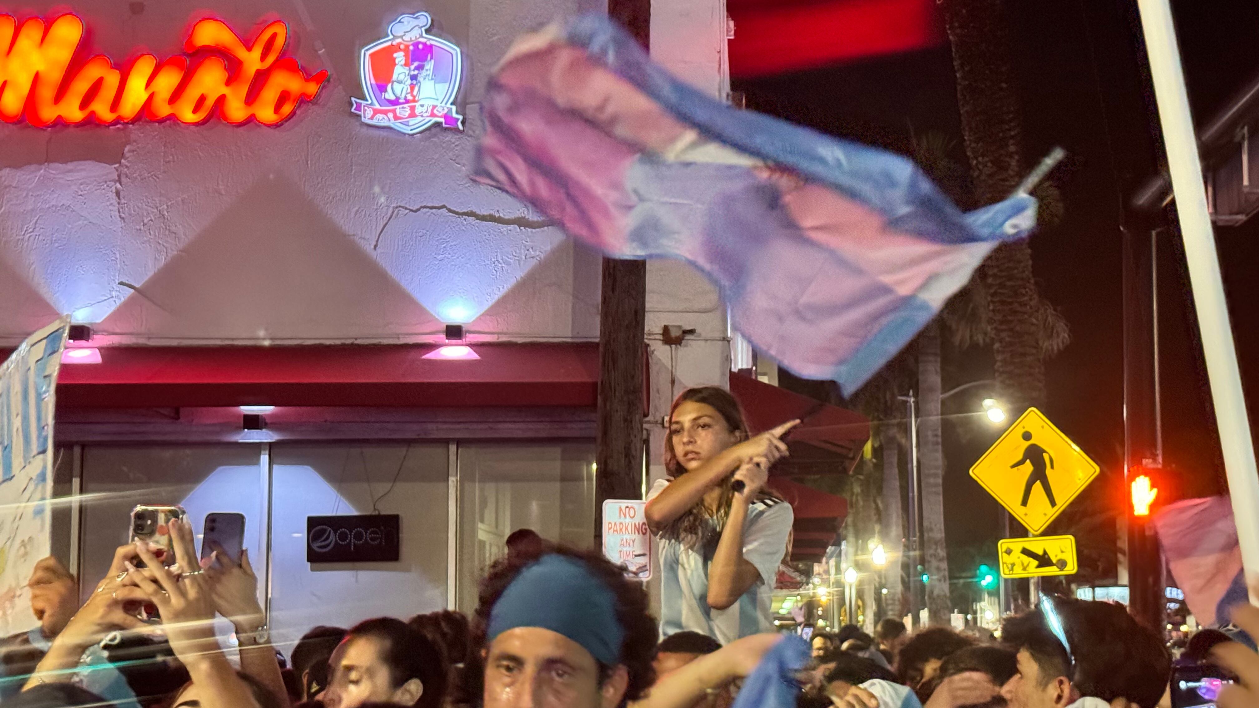 Una multitud de hinchas argentinos, vestidos con camisetas de la selección y agitando banderas celestes y blancas, celebra en las calles de Miami Beach durante la noche. La gente se muestra eufórica, tomando fotos y cantando, en una atmósfera festiva y llena de emoción tras la victoria de la Selección Argentina en la Copa América.