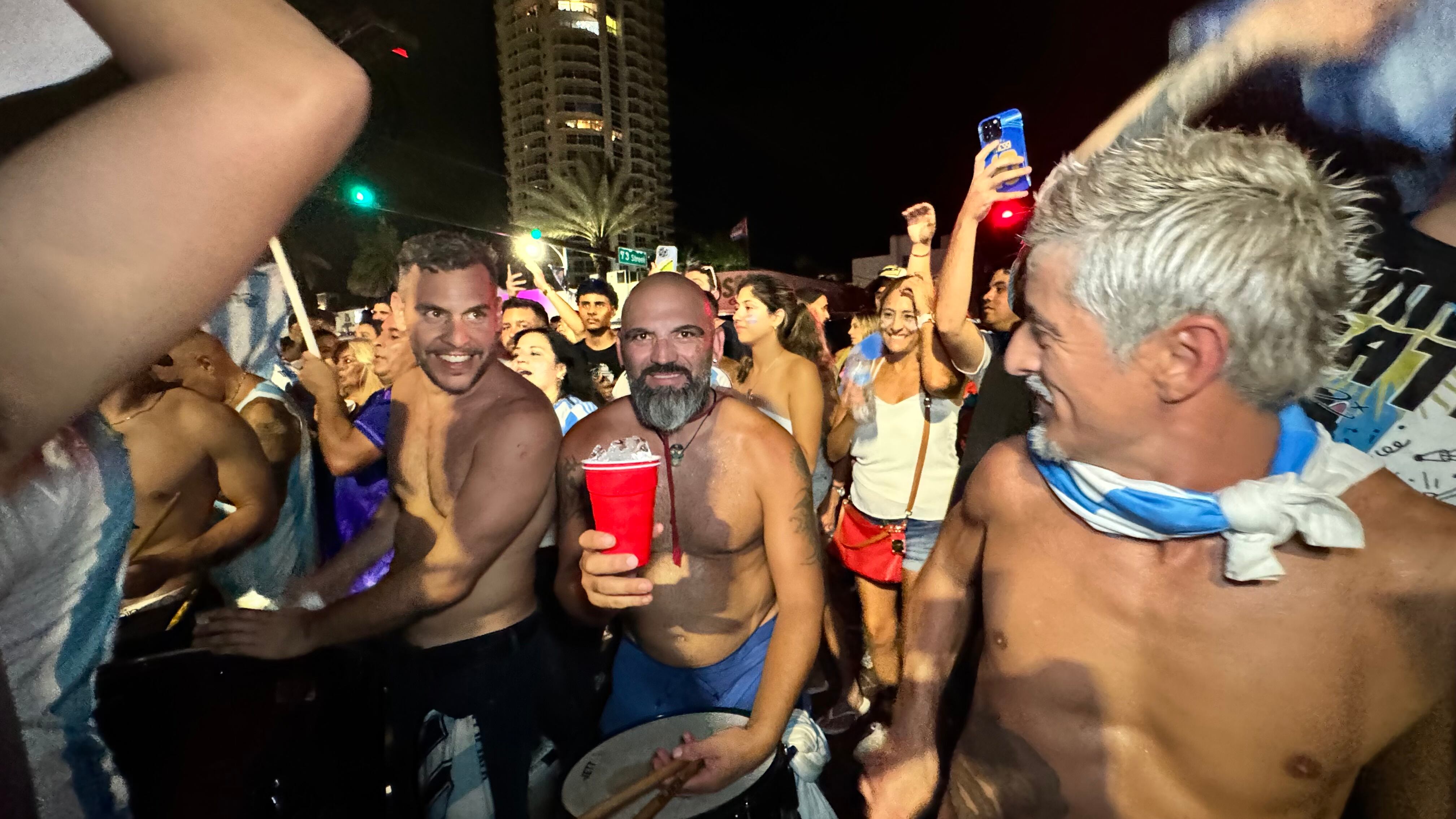 Una multitud de hinchas argentinos, vestidos con camisetas de la selección y agitando banderas celestes y blancas, celebra en las calles de Miami Beach durante la noche. La gente se muestra eufórica, tomando fotos y cantando, en una atmósfera festiva y llena de emoción tras la victoria de la Selección Argentina en la Copa América.