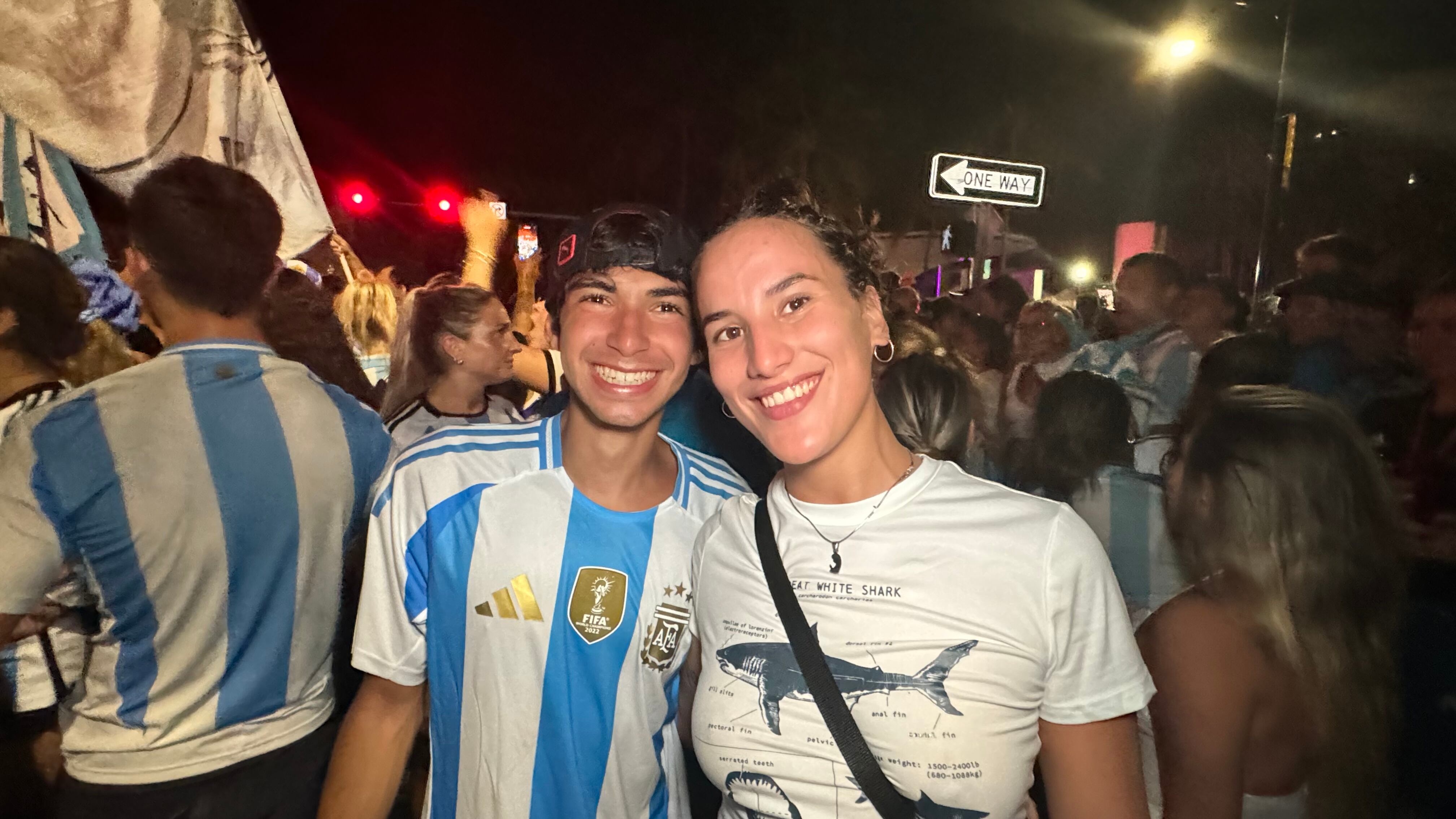 Una multitud de hinchas argentinos, vestidos con camisetas de la selección y agitando banderas celestes y blancas, celebra en las calles de Miami Beach durante la noche. La gente se muestra eufórica, tomando fotos y cantando, en una atmósfera festiva y llena de emoción tras la victoria de la Selección Argentina en la Copa América.