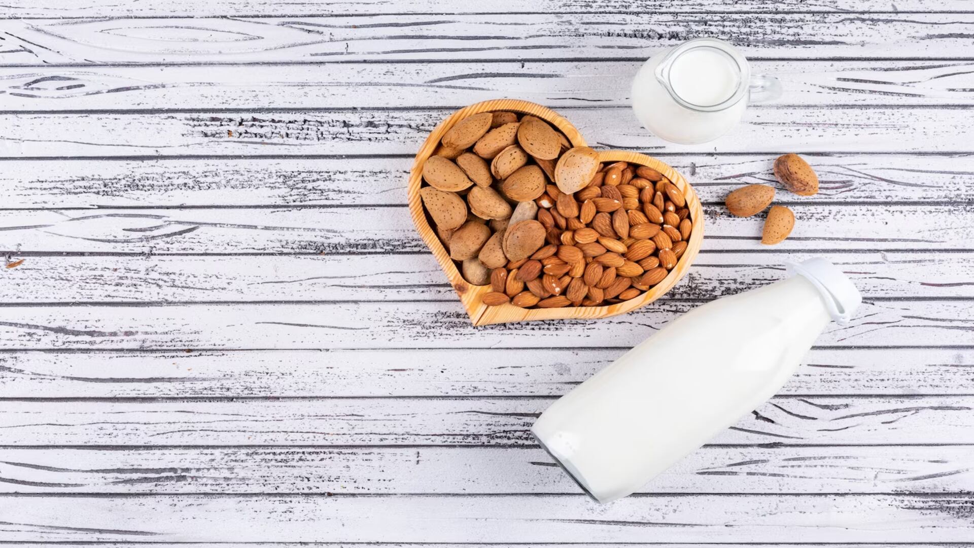 Almendra en plato demadera con forma de corazón y con botella de leche sobre mesa de madera blanca (Freepik)