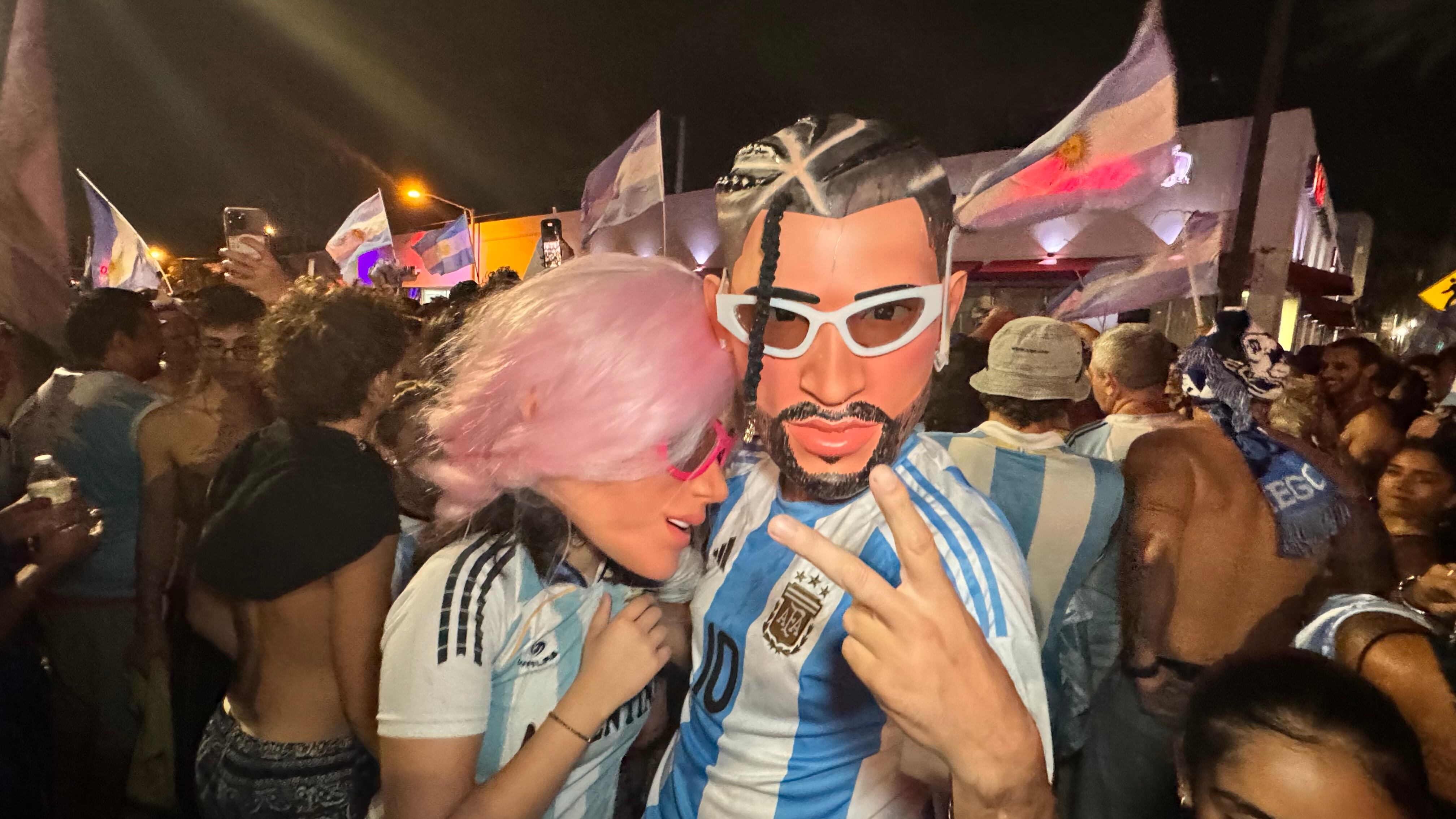 Una multitud de hinchas argentinos, vestidos con camisetas de la selección y agitando banderas celestes y blancas, celebra en las calles de Miami Beach durante la noche. La gente se muestra eufórica, tomando fotos y cantando, en una atmósfera festiva y llena de emoción tras la victoria de la Selección Argentina en la Copa América.