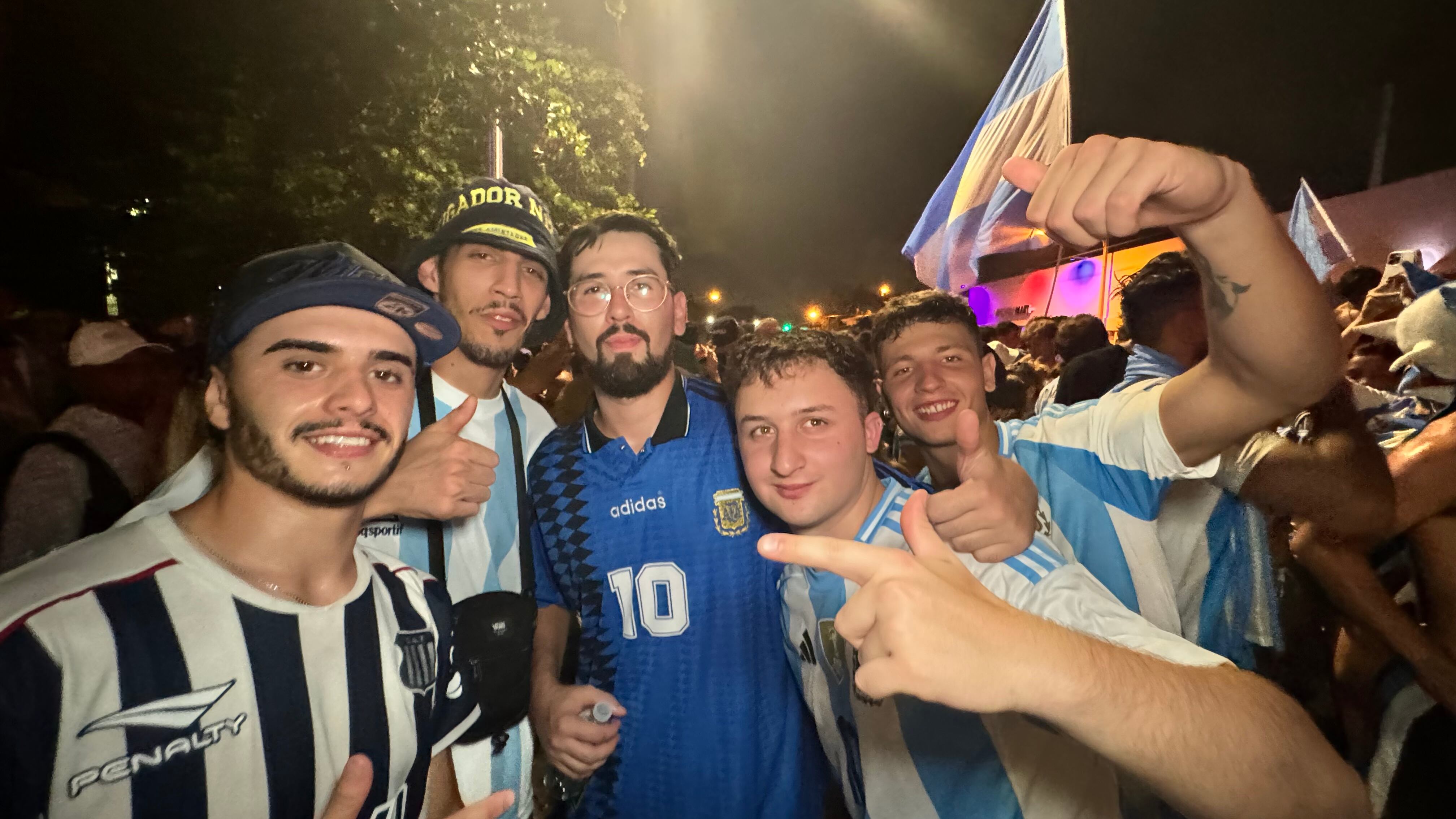 Una multitud de hinchas argentinos, vestidos con camisetas de la selección y agitando banderas celestes y blancas, celebra en las calles de Miami Beach durante la noche. La gente se muestra eufórica, tomando fotos y cantando, en una atmósfera festiva y llena de emoción tras la victoria de la Selección Argentina en la Copa América.