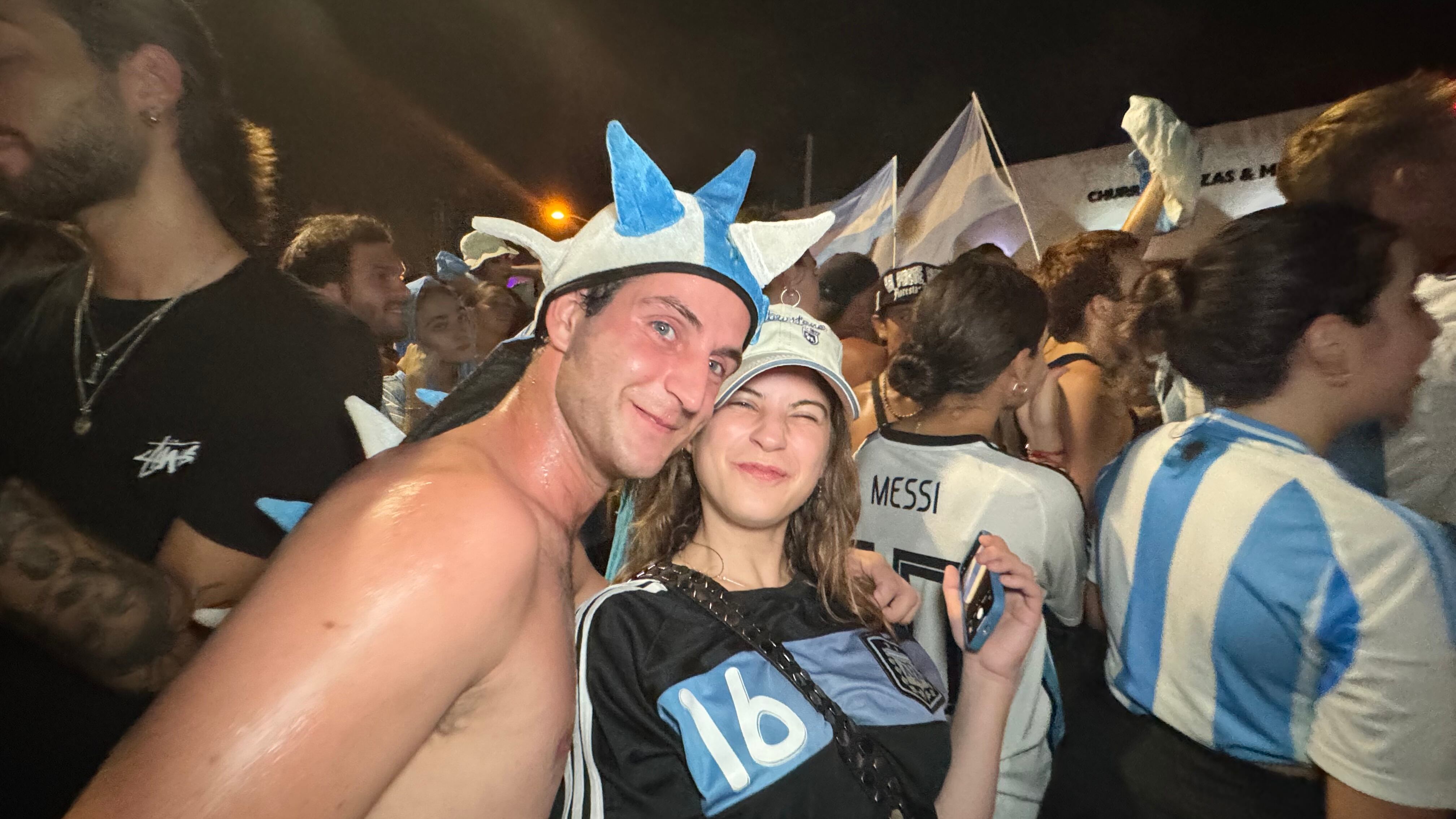 Una multitud de hinchas argentinos, vestidos con camisetas de la selección y agitando banderas celestes y blancas, celebra en las calles de Miami Beach durante la noche. La gente se muestra eufórica, tomando fotos y cantando, en una atmósfera festiva y llena de emoción tras la victoria de la Selección Argentina en la Copa América.