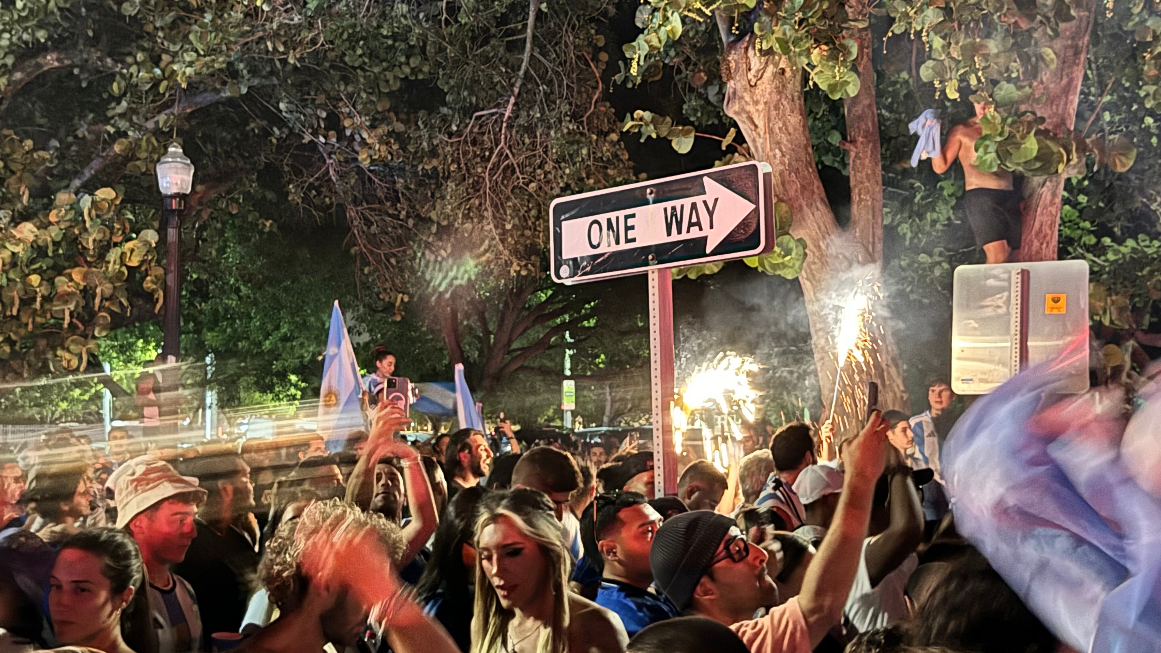 Una multitud de hinchas argentinos, vestidos con camisetas de la selección y agitando banderas celestes y blancas, celebra en las calles de Miami Beach durante la noche. La gente se muestra eufórica, tomando fotos y cantando, en una atmósfera festiva y llena de emoción tras la victoria de la Selección Argentina en la Copa América.