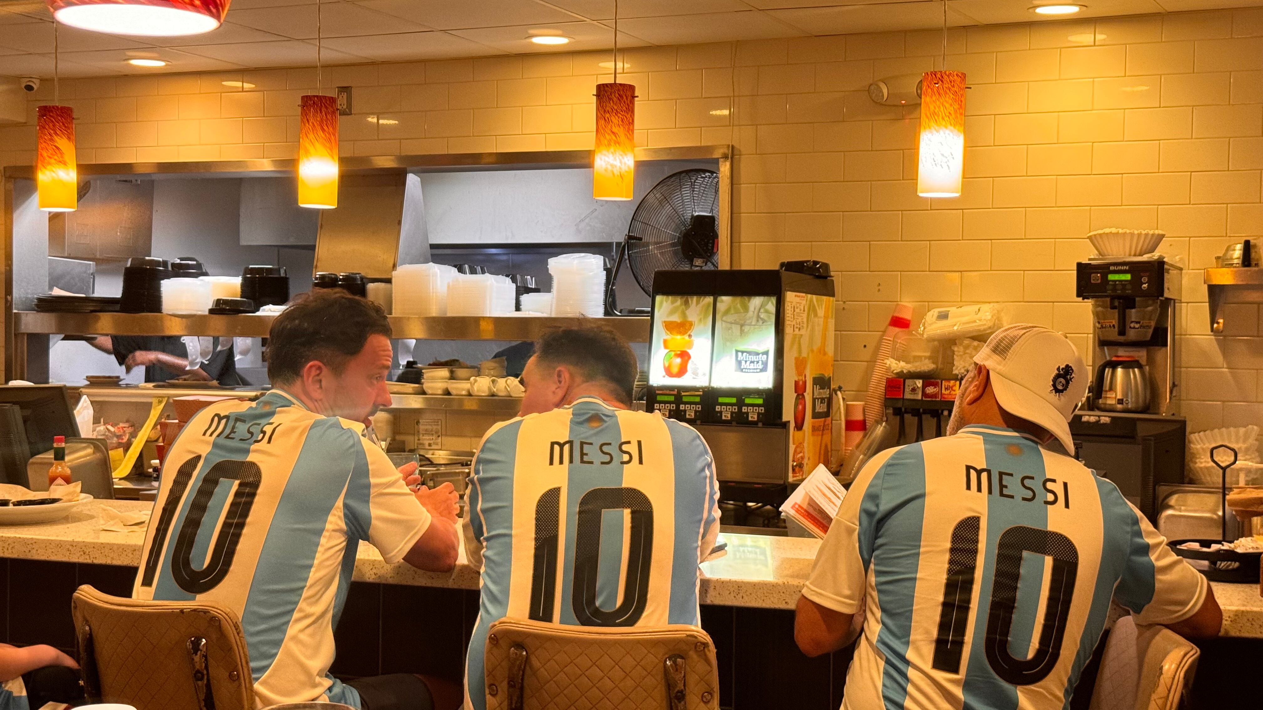 Una multitud de hinchas argentinos, vestidos con camisetas de la selección y agitando banderas celestes y blancas, celebra en las calles de Miami Beach durante la noche. La gente se muestra eufórica, tomando fotos y cantando, en una atmósfera festiva y llena de emoción tras la victoria de la Selección Argentina en la Copa América.