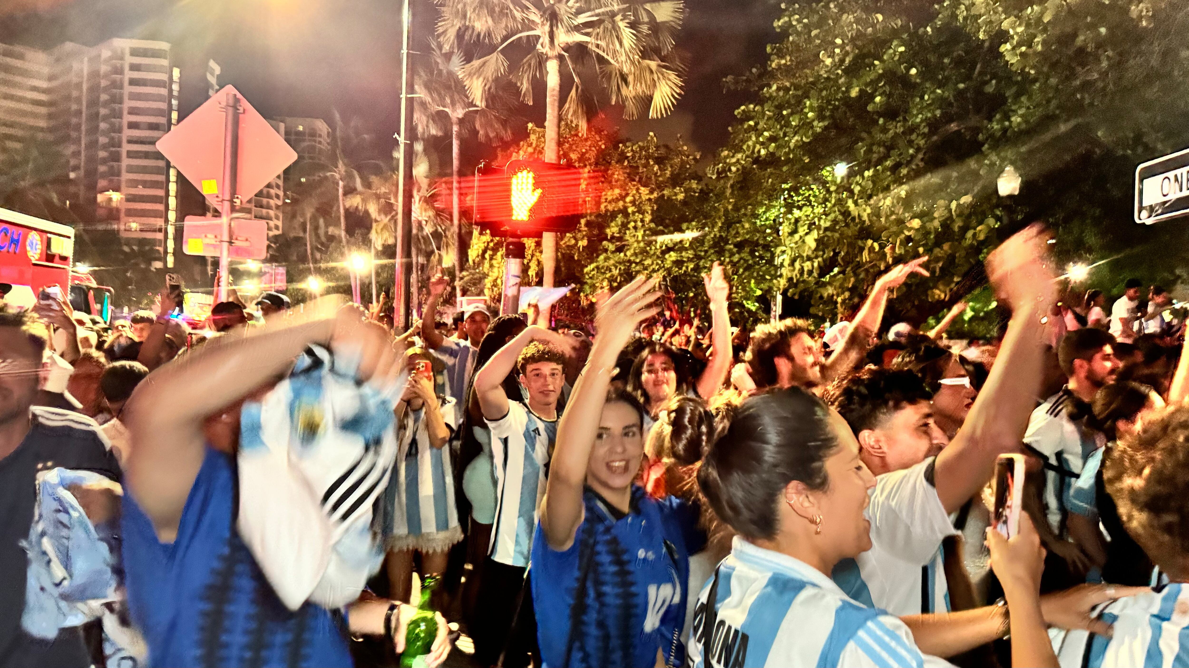 Una multitud de hinchas argentinos, vestidos con camisetas de la selección y agitando banderas celestes y blancas, celebra en las calles de Miami Beach durante la noche. La gente se muestra eufórica, tomando fotos y cantando, en una atmósfera festiva y llena de emoción tras la victoria de la Selección Argentina en la Copa América.