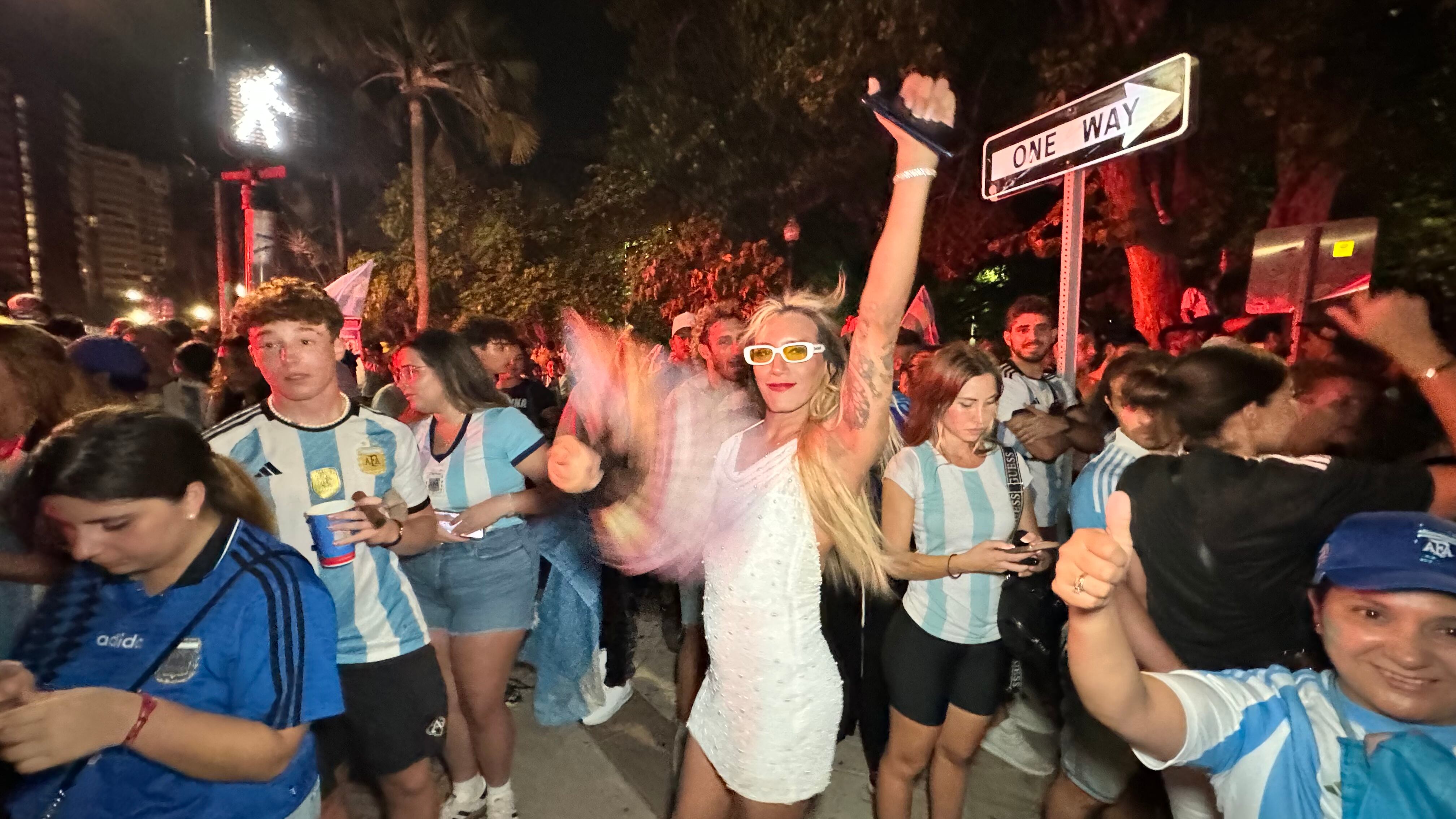 Una multitud de hinchas argentinos, vestidos con camisetas de la selección y agitando banderas celestes y blancas, celebra en las calles de Miami Beach durante la noche. La gente se muestra eufórica, tomando fotos y cantando, en una atmósfera festiva y llena de emoción tras la victoria de la Selección Argentina en la Copa América.