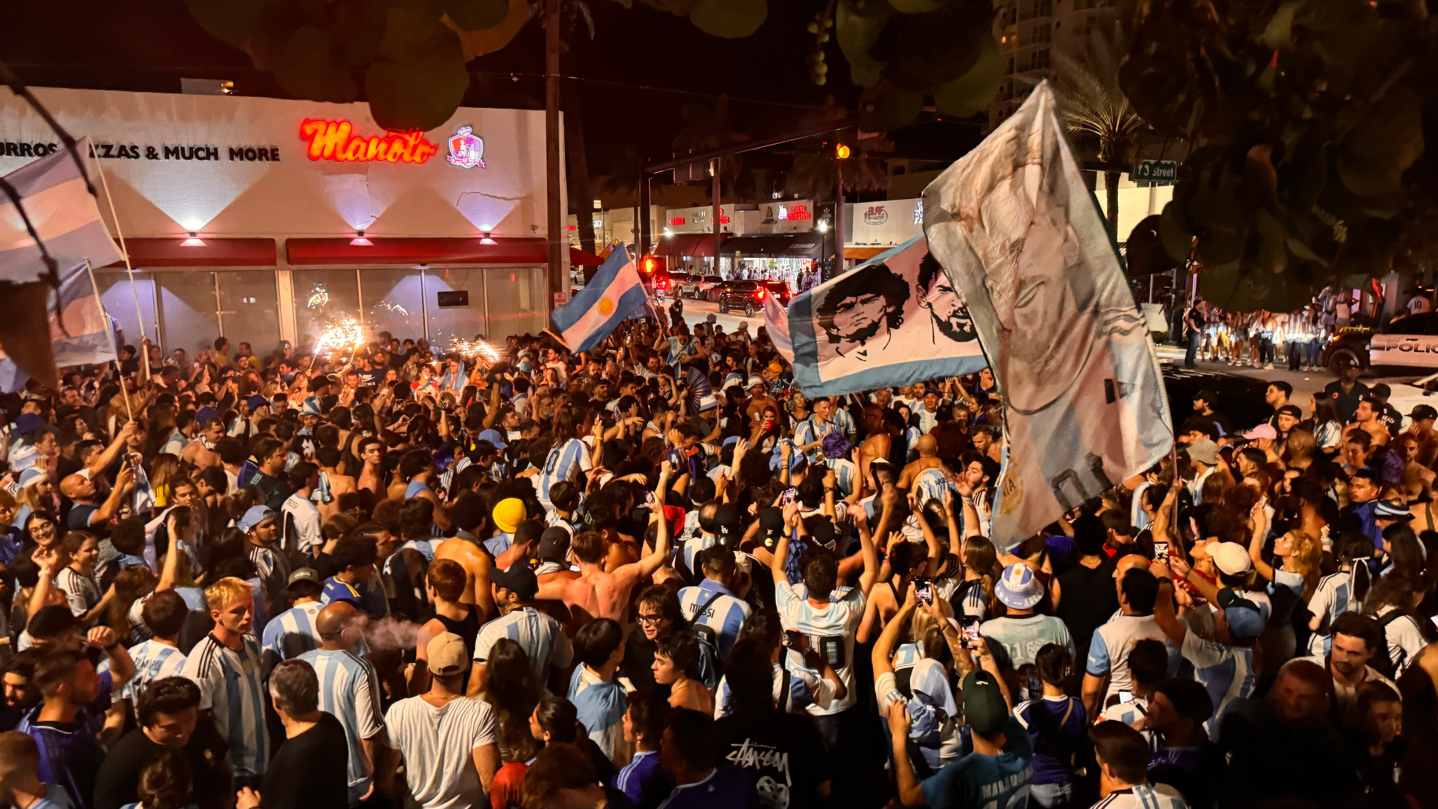 Una multitud de hinchas argentinos, vestidos con camisetas de la selección y agitando banderas celestes y blancas, celebra en las calles de Miami Beach durante la noche. La gente se muestra eufórica, tomando fotos y cantando, en una atmósfera festiva y llena de emoción tras la victoria de la Selección Argentina en la Copa América.