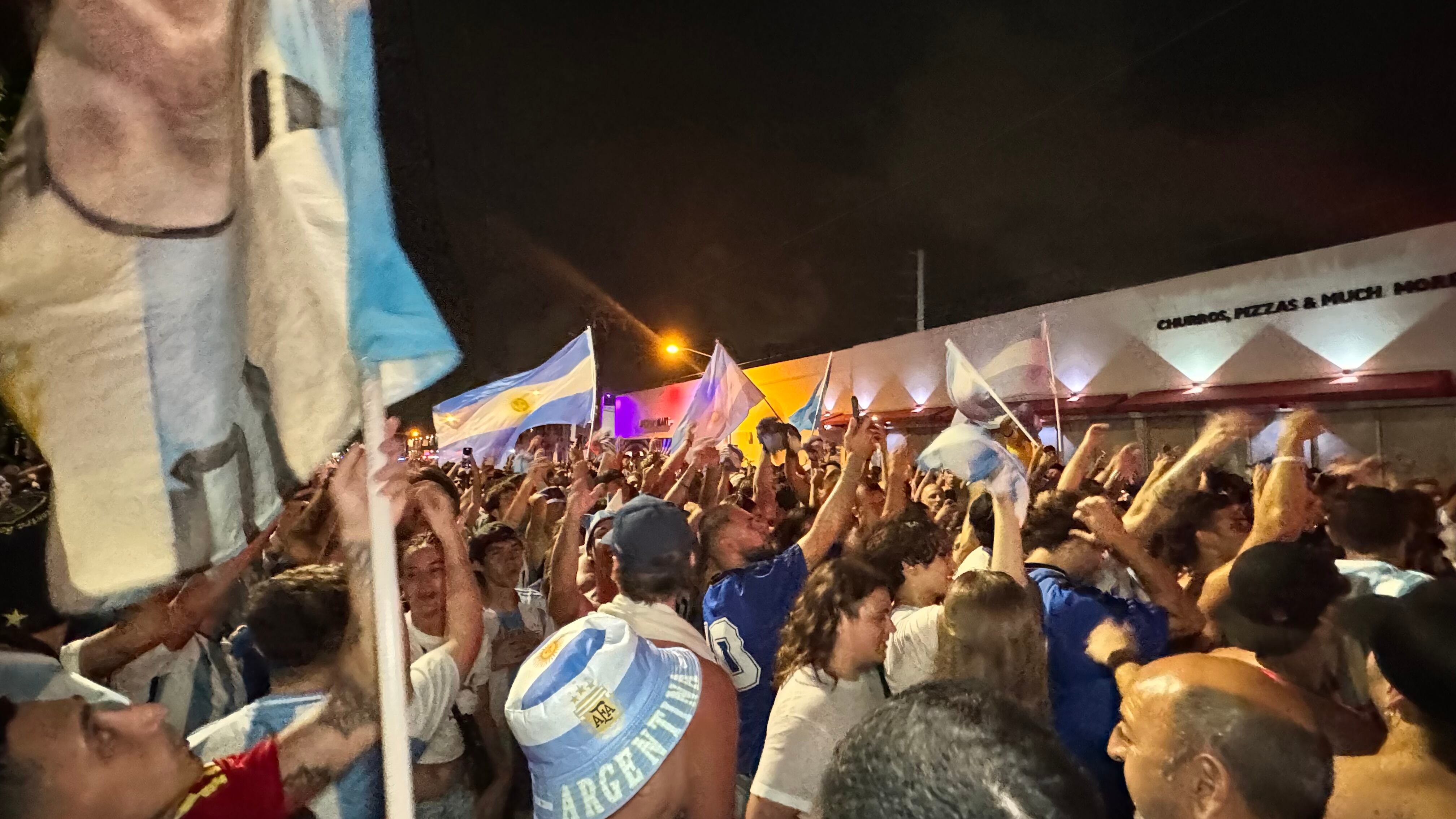 Una multitud de hinchas argentinos, vestidos con camisetas de la selección y agitando banderas celestes y blancas, celebra en las calles de Miami Beach durante la noche. La gente se muestra eufórica, tomando fotos y cantando, en una atmósfera festiva y llena de emoción tras la victoria de la Selección Argentina en la Copa América.