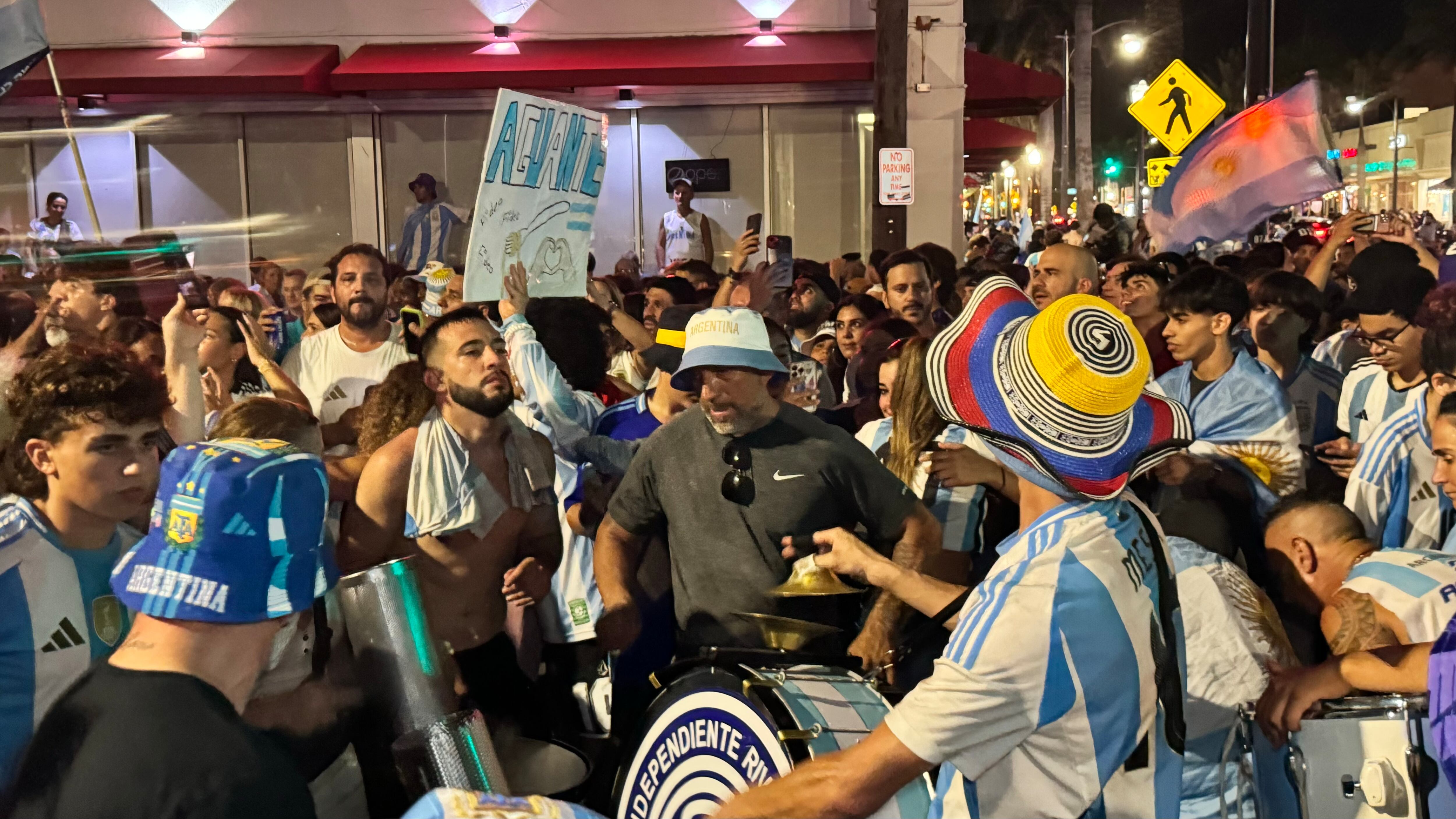 Una multitud de hinchas argentinos, vestidos con camisetas de la selección y agitando banderas celestes y blancas, celebra en las calles de Miami Beach durante la noche. La gente se muestra eufórica, tomando fotos y cantando, en una atmósfera festiva y llena de emoción tras la victoria de la Selección Argentina en la Copa América.