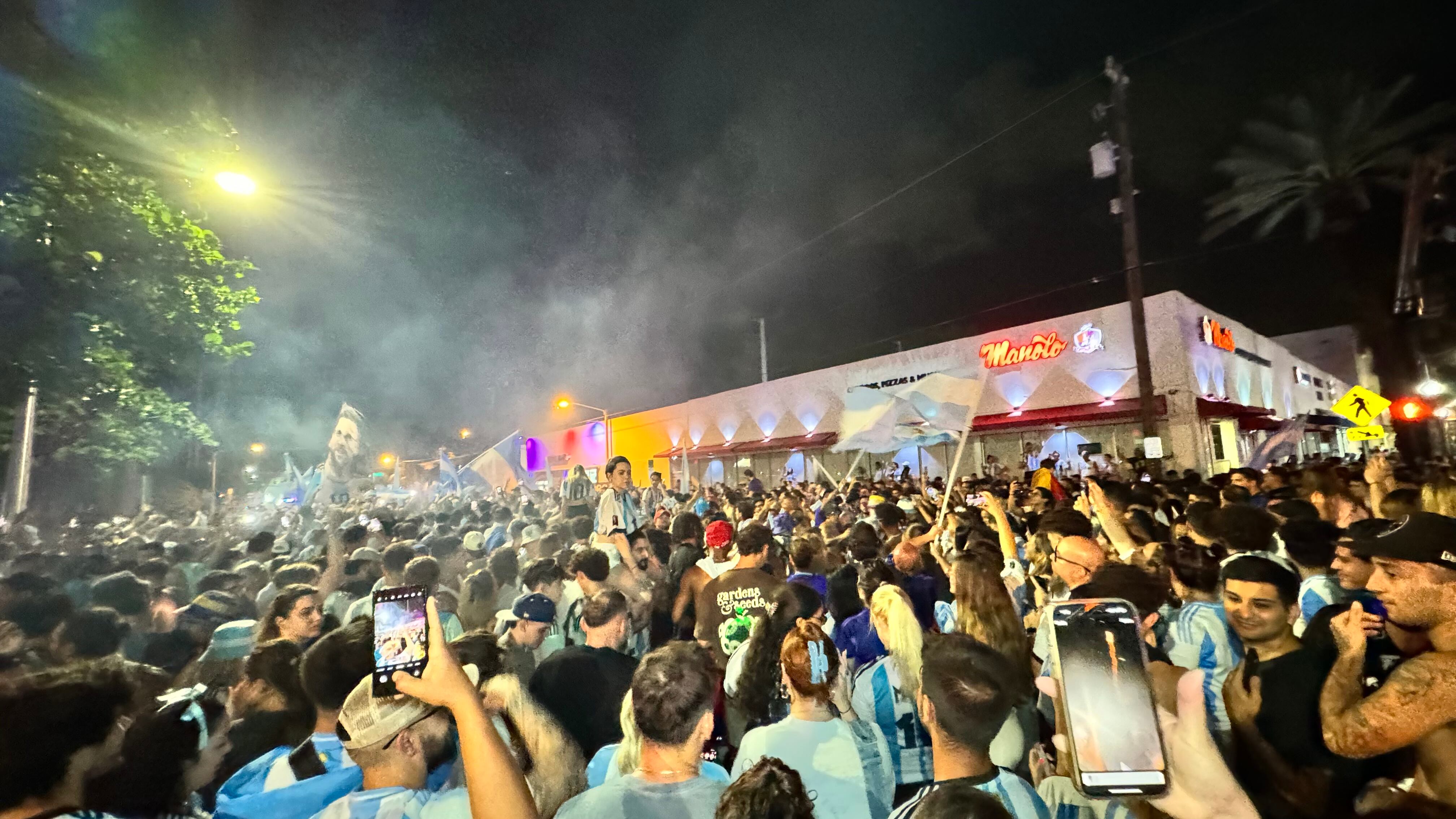 Una multitud de hinchas argentinos, vestidos con camisetas de la selección y agitando banderas celestes y blancas, celebra en las calles de Miami Beach durante la noche. La gente se muestra eufórica, tomando fotos y cantando, en una atmósfera festiva y llena de emoción tras la victoria de la Selección Argentina en la Copa América.