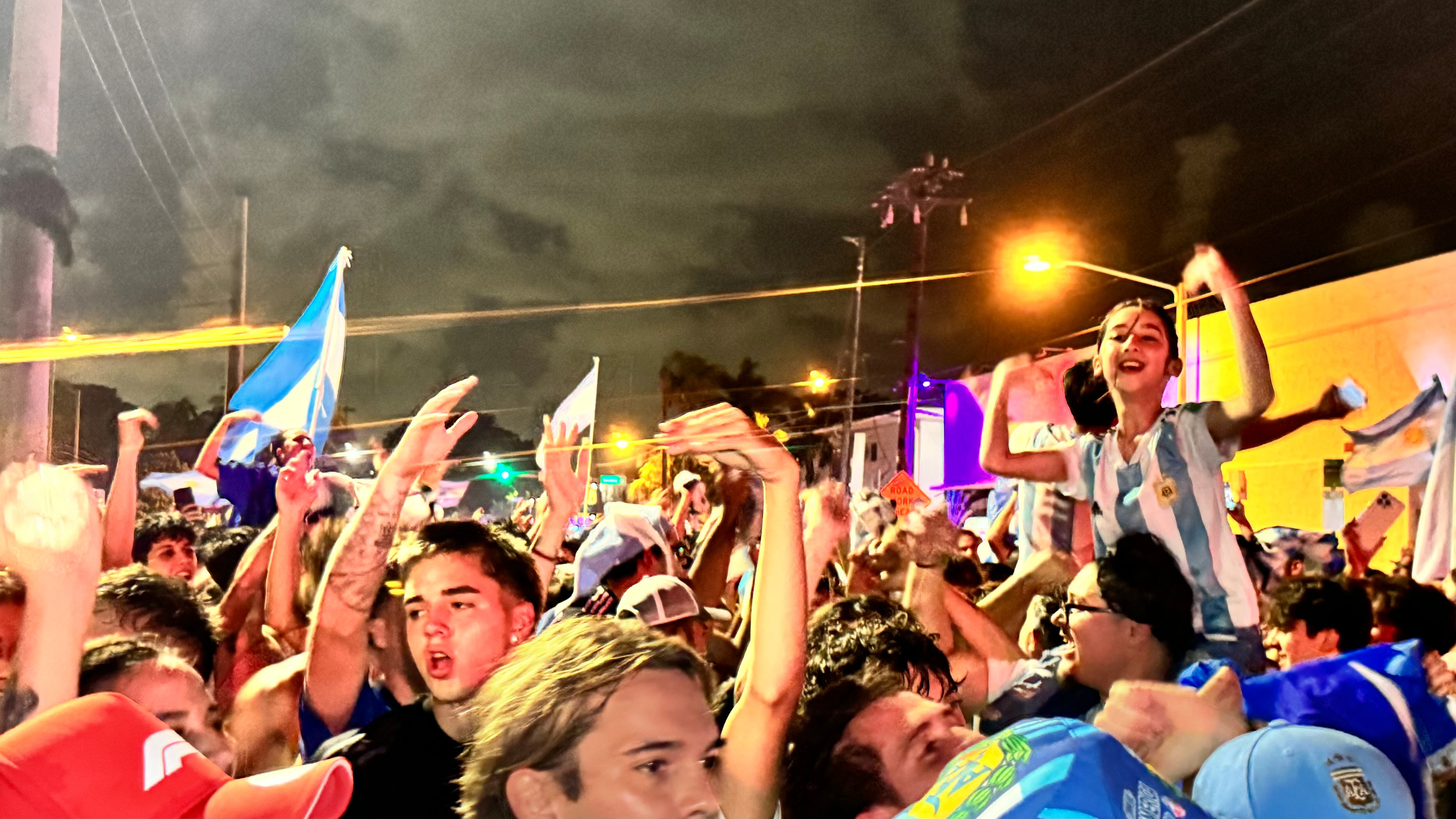 Una multitud de hinchas argentinos, vestidos con camisetas de la selección y agitando banderas celestes y blancas, celebra en las calles de Miami Beach durante la noche. La gente se muestra eufórica, tomando fotos y cantando, en una atmósfera festiva y llena de emoción tras la victoria de la Selección Argentina en la Copa América.