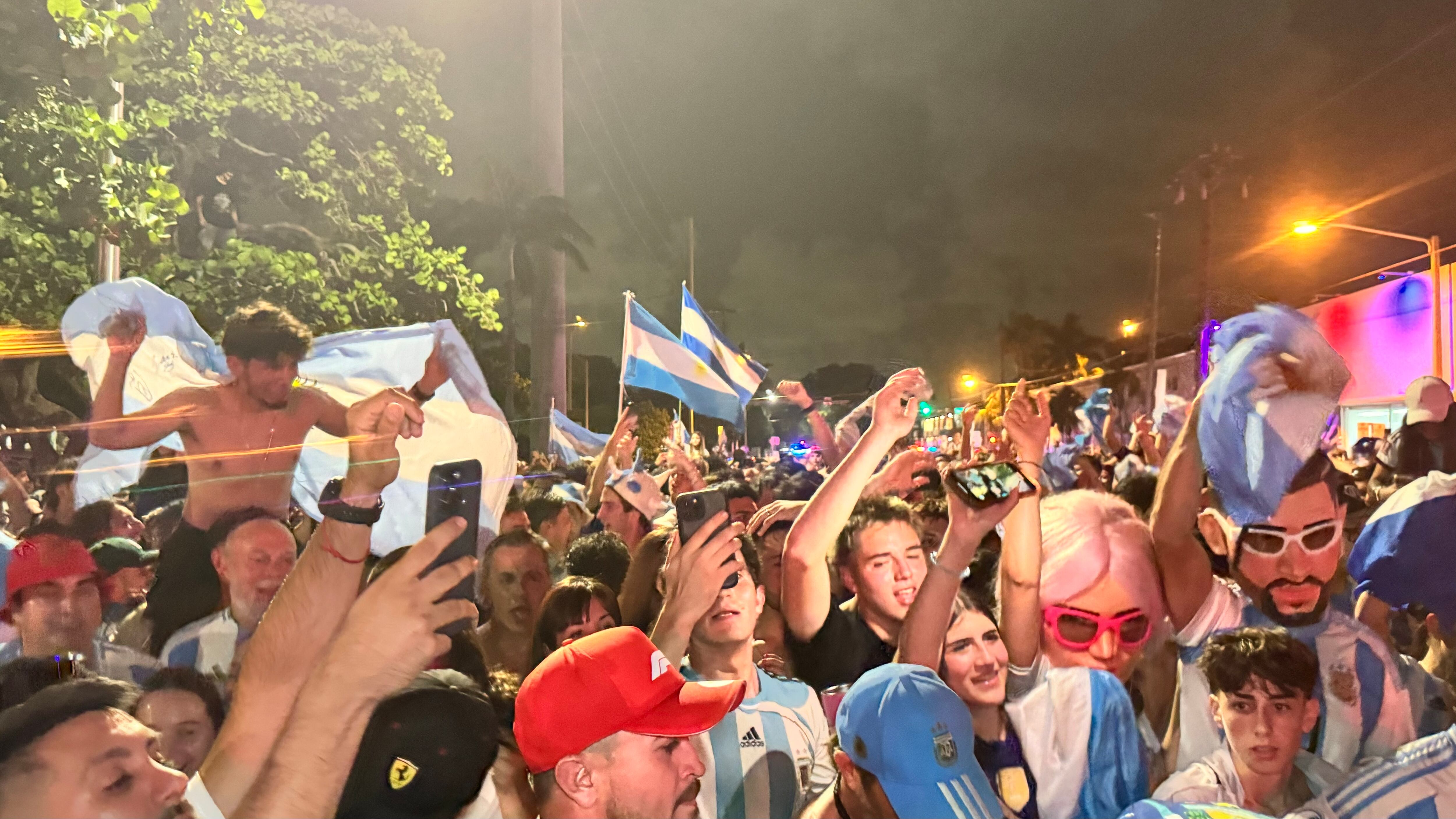 Una multitud de hinchas argentinos, vestidos con camisetas de la selección y agitando banderas celestes y blancas, celebra en las calles de Miami Beach durante la noche. La gente se muestra eufórica, tomando fotos y cantando, en una atmósfera festiva y llena de emoción tras la victoria de la Selección Argentina en la Copa América.