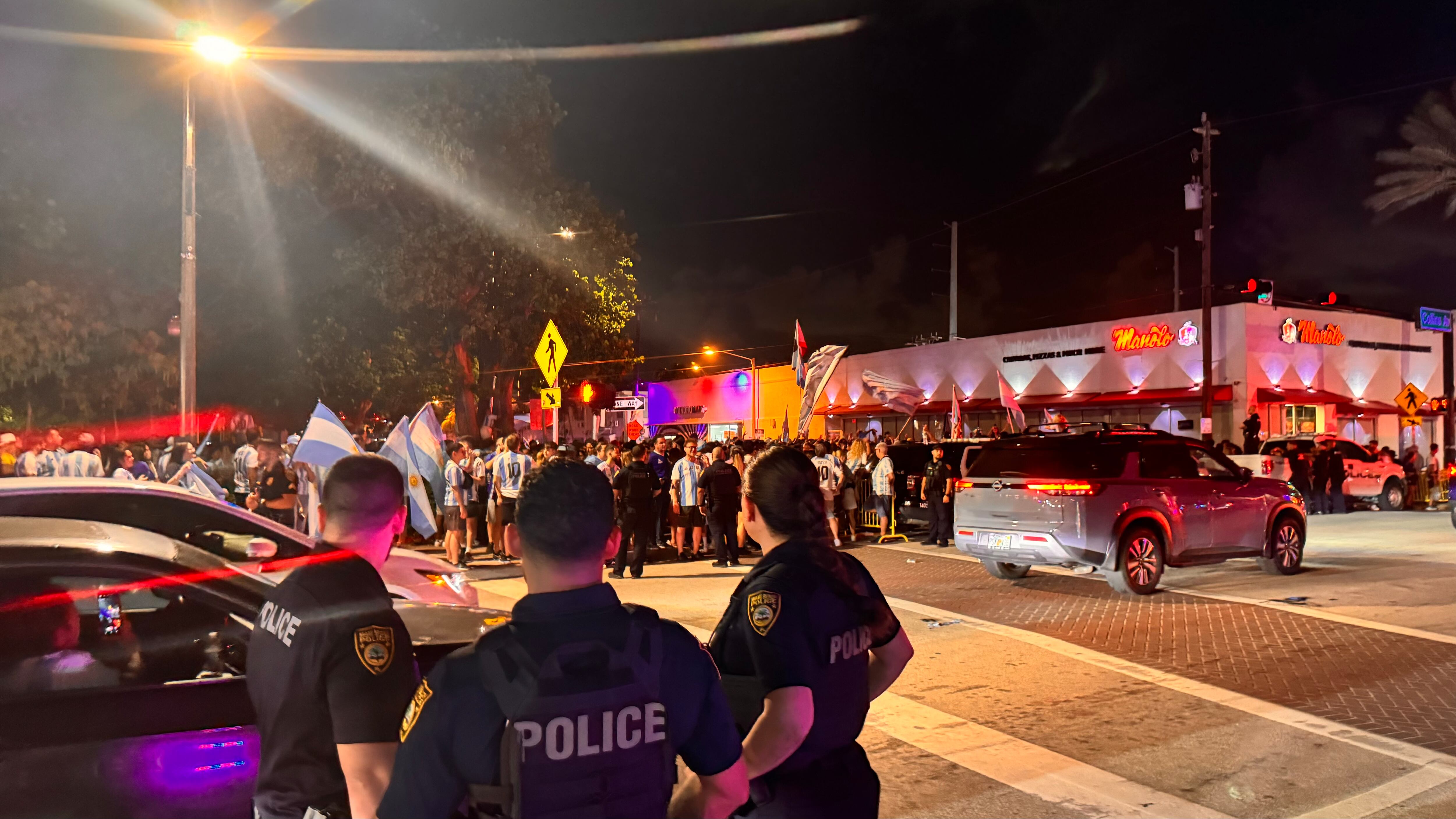 Una multitud de hinchas argentinos, vestidos con camisetas de la selección y agitando banderas celestes y blancas, celebra en las calles de Miami Beach durante la noche. La gente se muestra eufórica, tomando fotos y cantando, en una atmósfera festiva y llena de emoción tras la victoria de la Selección Argentina en la Copa América.