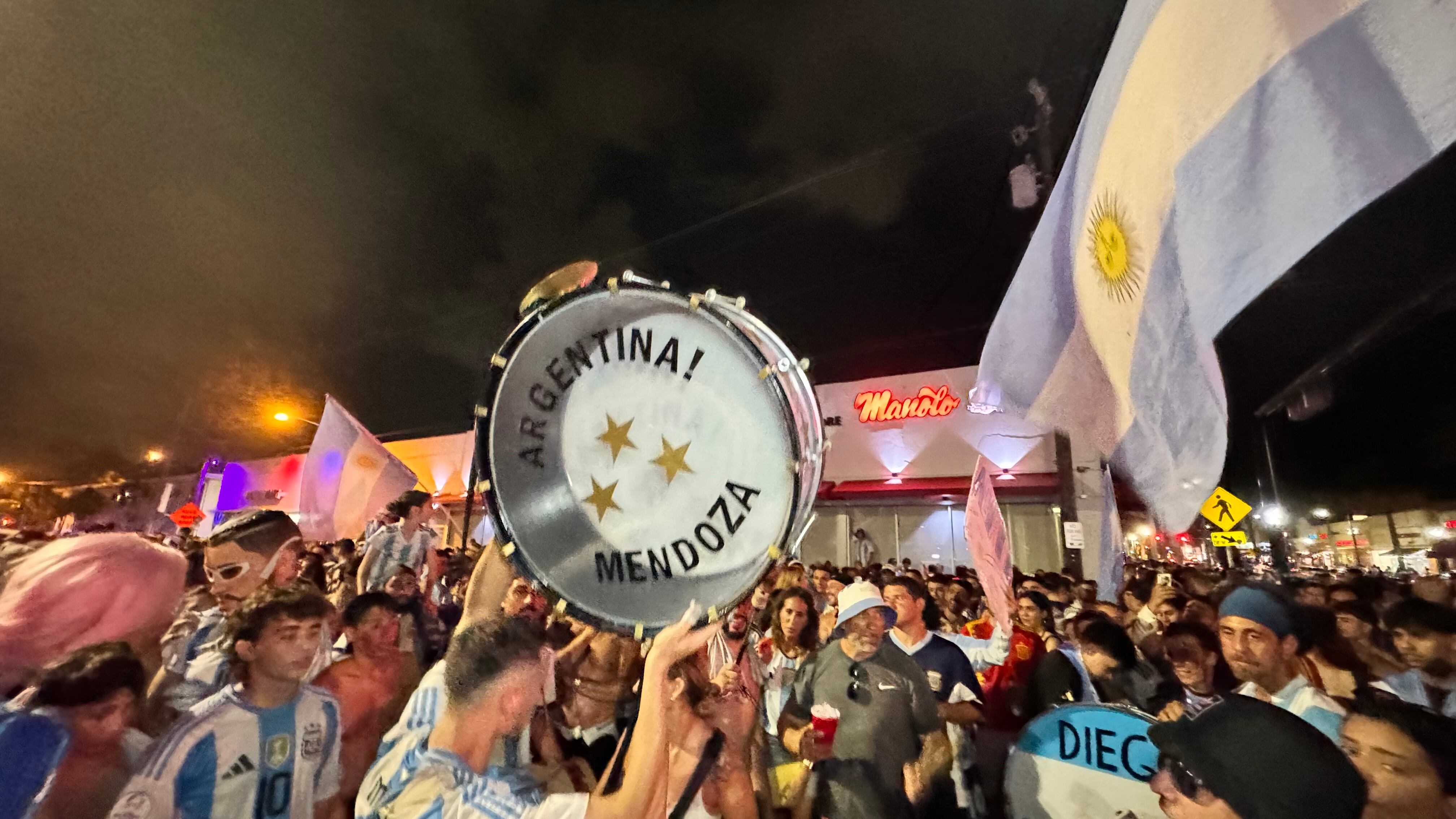 Una multitud de hinchas argentinos, vestidos con camisetas de la selección y agitando banderas celestes y blancas, celebra en las calles de Miami Beach durante la noche. La gente se muestra eufórica, tomando fotos y cantando, en una atmósfera festiva y llena de emoción tras la victoria de la Selección Argentina en la Copa América.