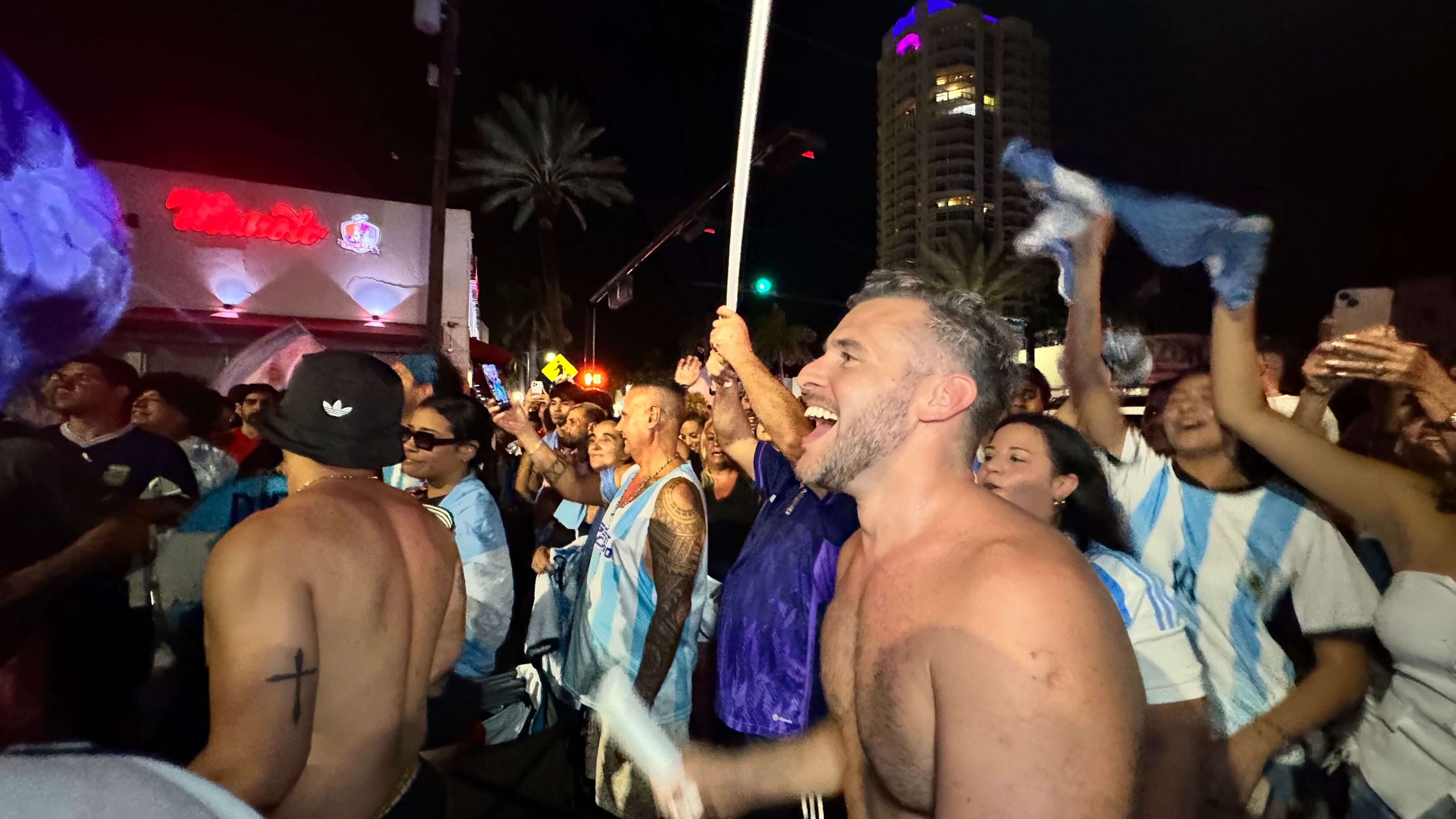 Una multitud de hinchas argentinos, vestidos con camisetas de la selección y agitando banderas celestes y blancas, celebra en las calles de Miami Beach durante la noche. La gente se muestra eufórica, tomando fotos y cantando, en una atmósfera festiva y llena de emoción tras la victoria de la Selección Argentina en la Copa América.