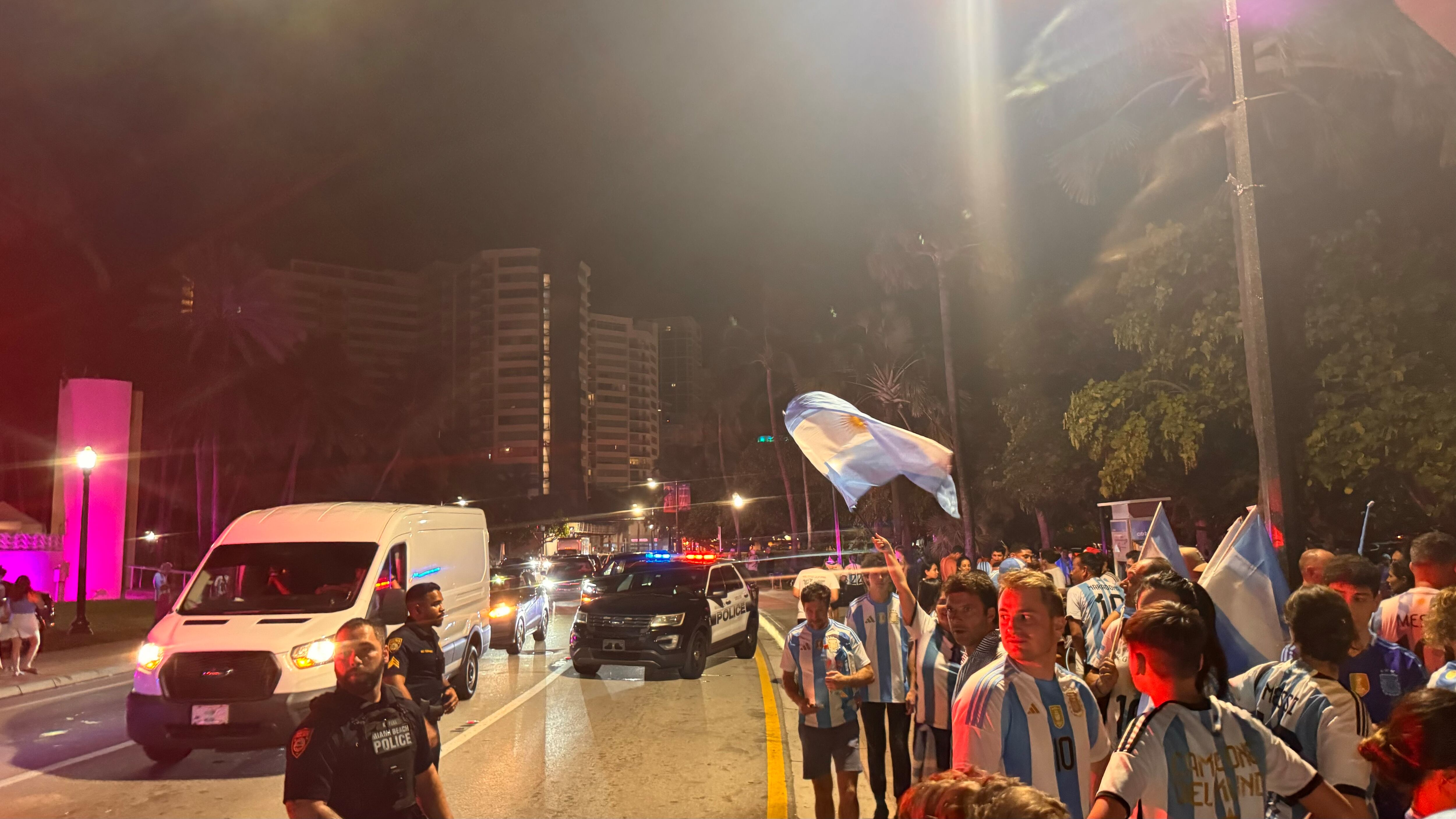 Una multitud de hinchas argentinos, vestidos con camisetas de la selección y agitando banderas celestes y blancas, celebra en las calles de Miami Beach durante la noche. La gente se muestra eufórica, tomando fotos y cantando, en una atmósfera festiva y llena de emoción tras la victoria de la Selección Argentina en la Copa América.