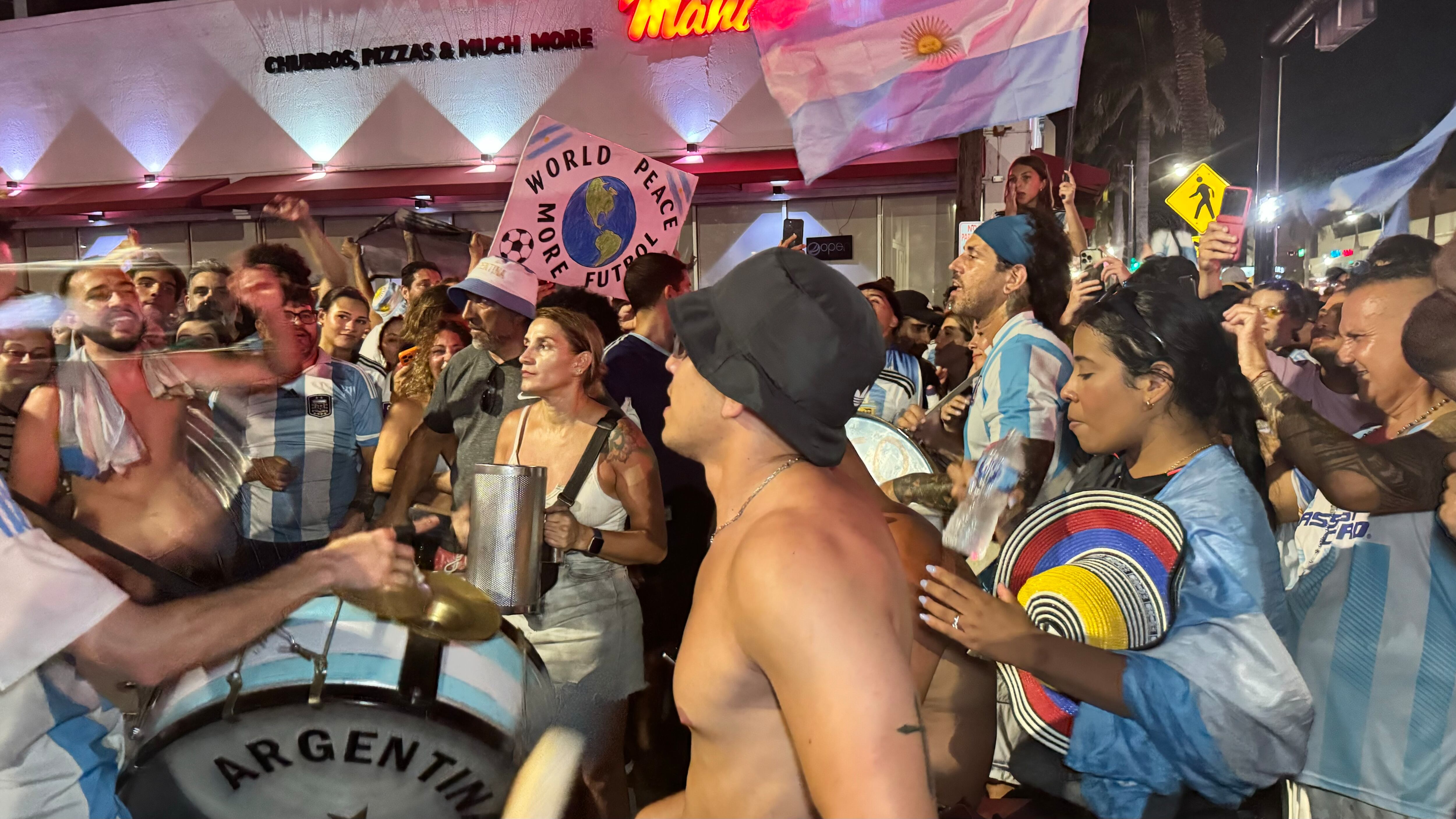 Una multitud de hinchas argentinos, vestidos con camisetas de la selección y agitando banderas celestes y blancas, celebra en las calles de Miami Beach durante la noche. La gente se muestra eufórica, tomando fotos y cantando, en una atmósfera festiva y llena de emoción tras la victoria de la Selección Argentina en la Copa América.