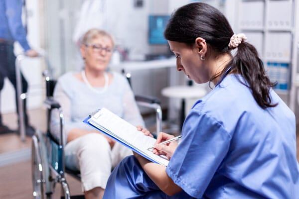 Una doctora escribiendo un informe mientras atienda a una paciente de Esclerosis Lateral Amiotrófica - ELA en silla de ruedas (Yeyehelp)