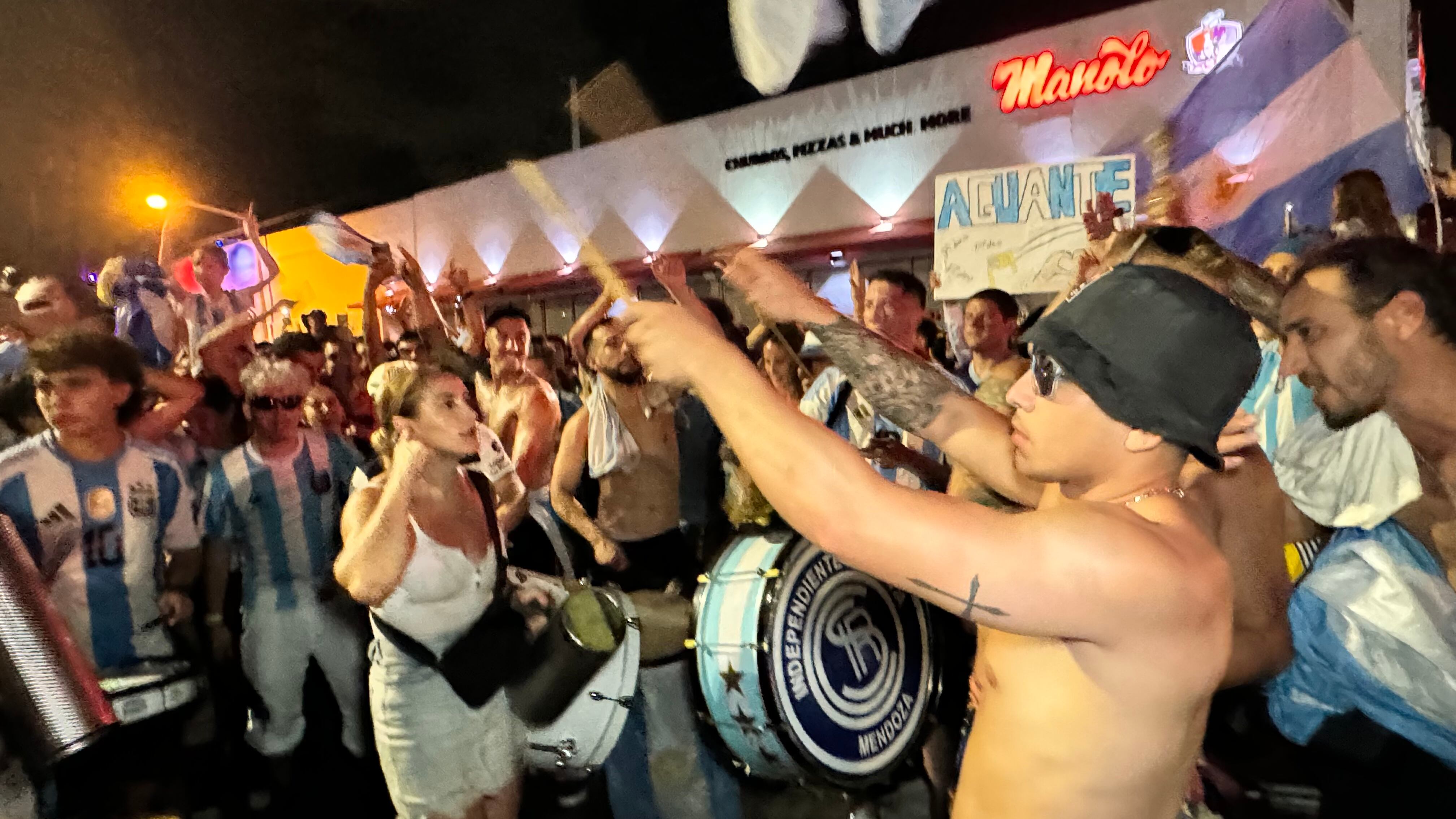 Una multitud de hinchas argentinos, vestidos con camisetas de la selección y agitando banderas celestes y blancas, celebra en las calles de Miami Beach durante la noche. La gente se muestra eufórica, tomando fotos y cantando, en una atmósfera festiva y llena de emoción tras la victoria de la Selección Argentina en la Copa América.