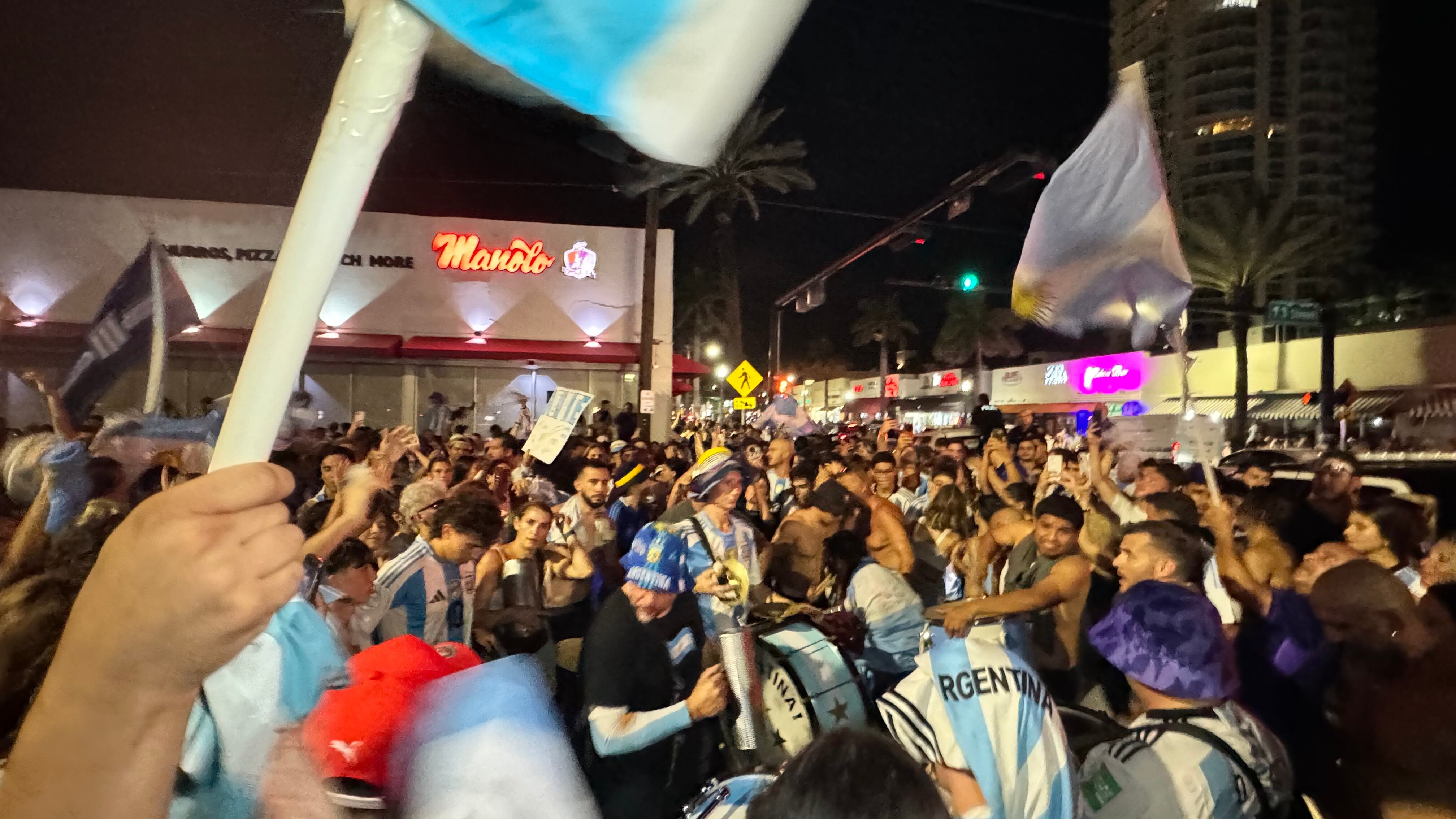 Una multitud de hinchas argentinos, vestidos con camisetas de la selección y agitando banderas celestes y blancas, celebra en las calles de Miami Beach durante la noche. La gente se muestra eufórica, tomando fotos y cantando, en una atmósfera festiva y llena de emoción tras la victoria de la Selección Argentina en la Copa América.