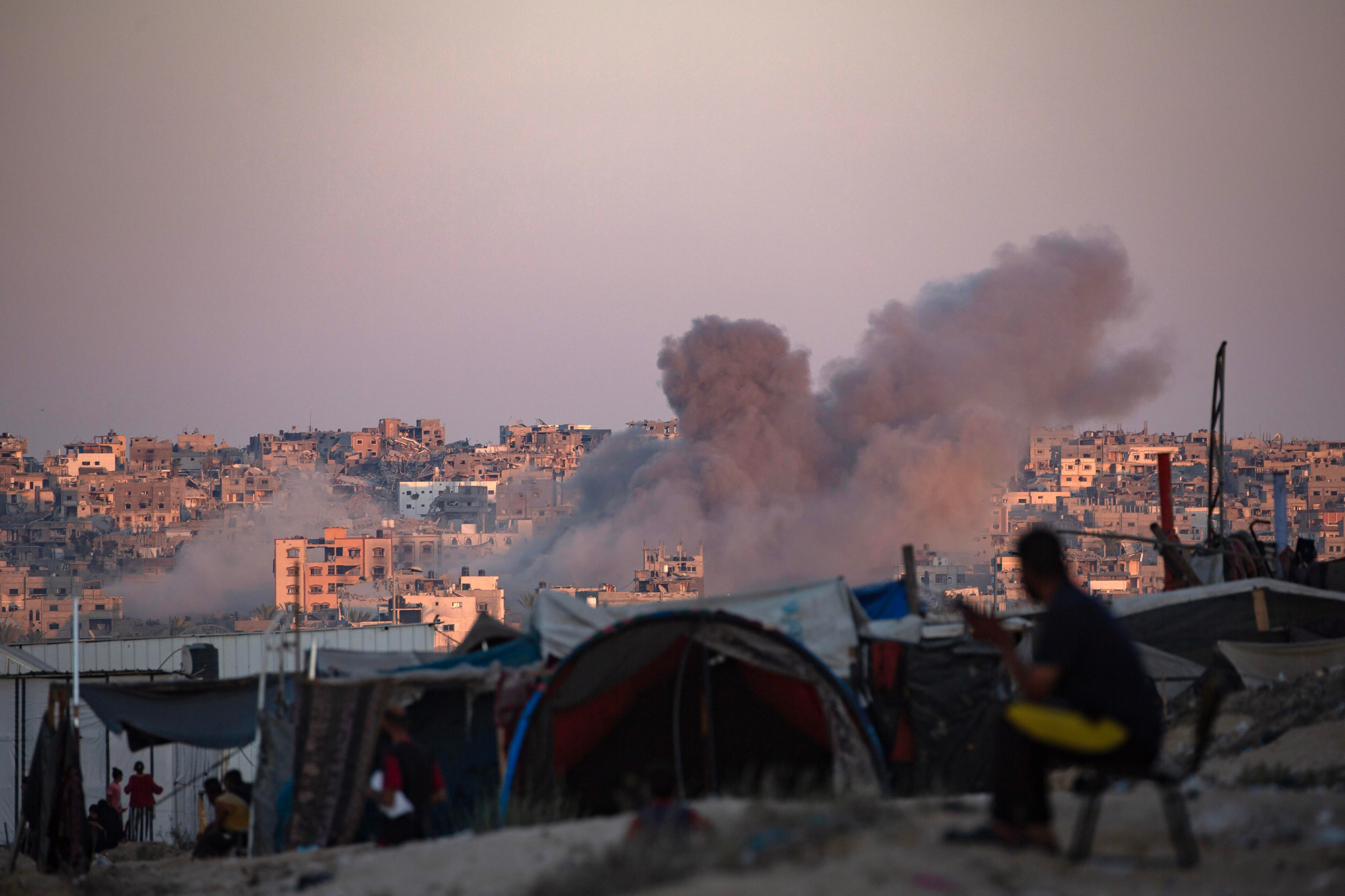 El humo se eleva tras un ataque aéreo israelí mientras los palestinos desplazados internos se sientan junto a sus tiendas de campaña en el campamento de Khan Younis, al sur de la Franja de Gaza (EFE/Haitram Imad)
