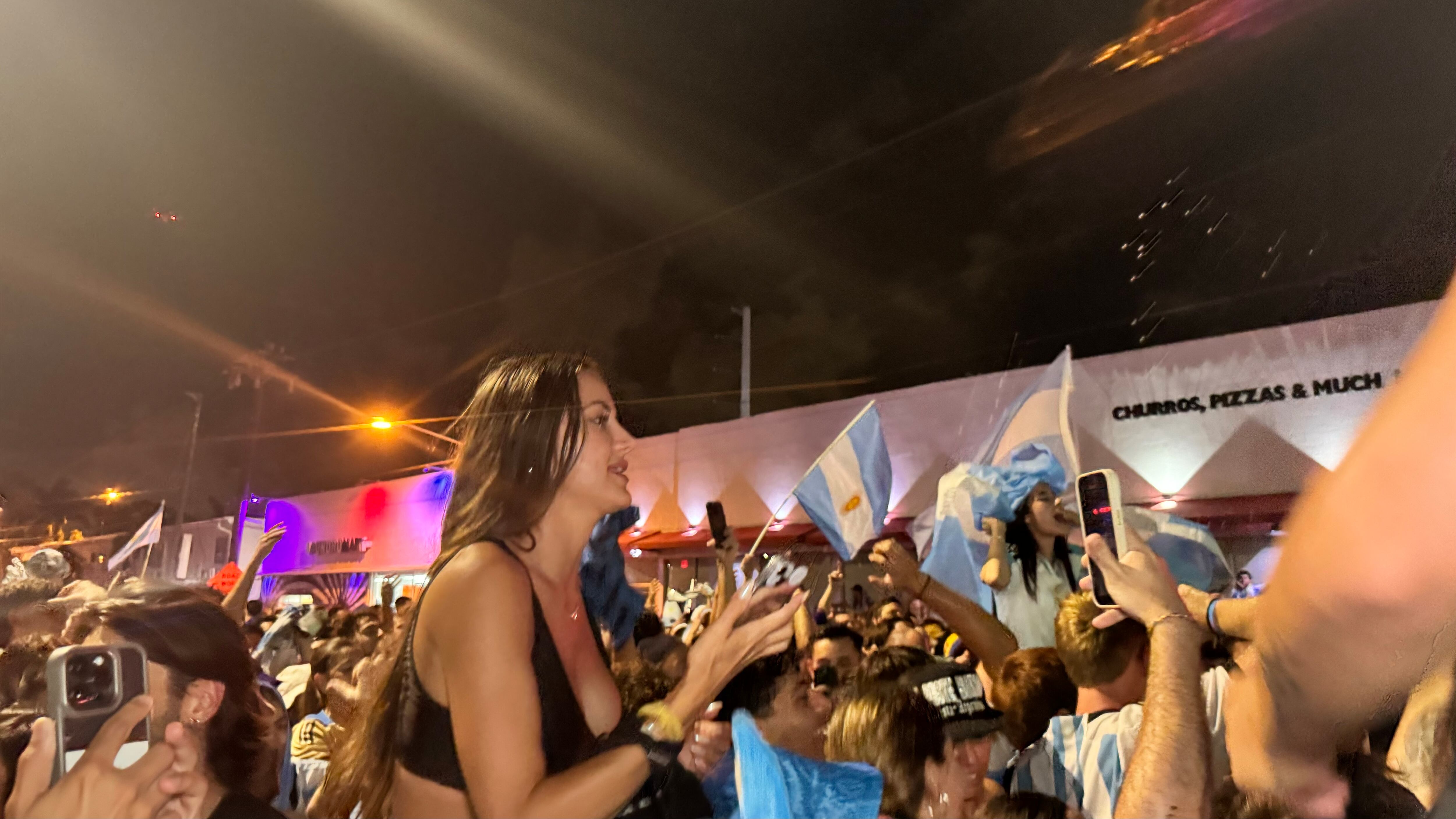 Una multitud de hinchas argentinos, vestidos con camisetas de la selección y agitando banderas celestes y blancas, celebra en las calles de Miami Beach durante la noche. La gente se muestra eufórica, tomando fotos y cantando, en una atmósfera festiva y llena de emoción tras la victoria de la Selección Argentina en la Copa América.