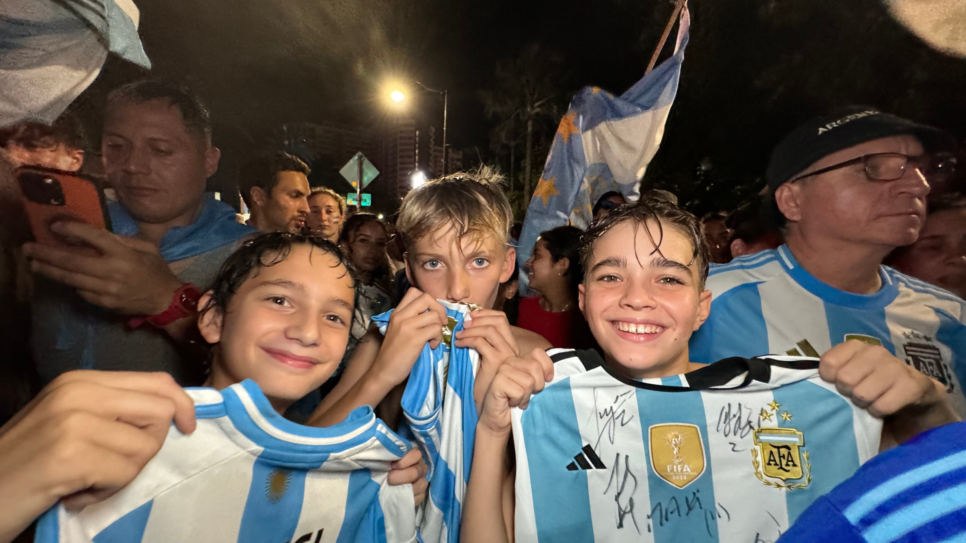 Una multitud de hinchas argentinos, vestidos con camisetas de la selección y agitando banderas celestes y blancas, celebra en las calles de Miami Beach durante la noche. La gente se muestra eufórica, tomando fotos y cantando, en una atmósfera festiva y llena de emoción tras la victoria de la Selección Argentina en la Copa América.